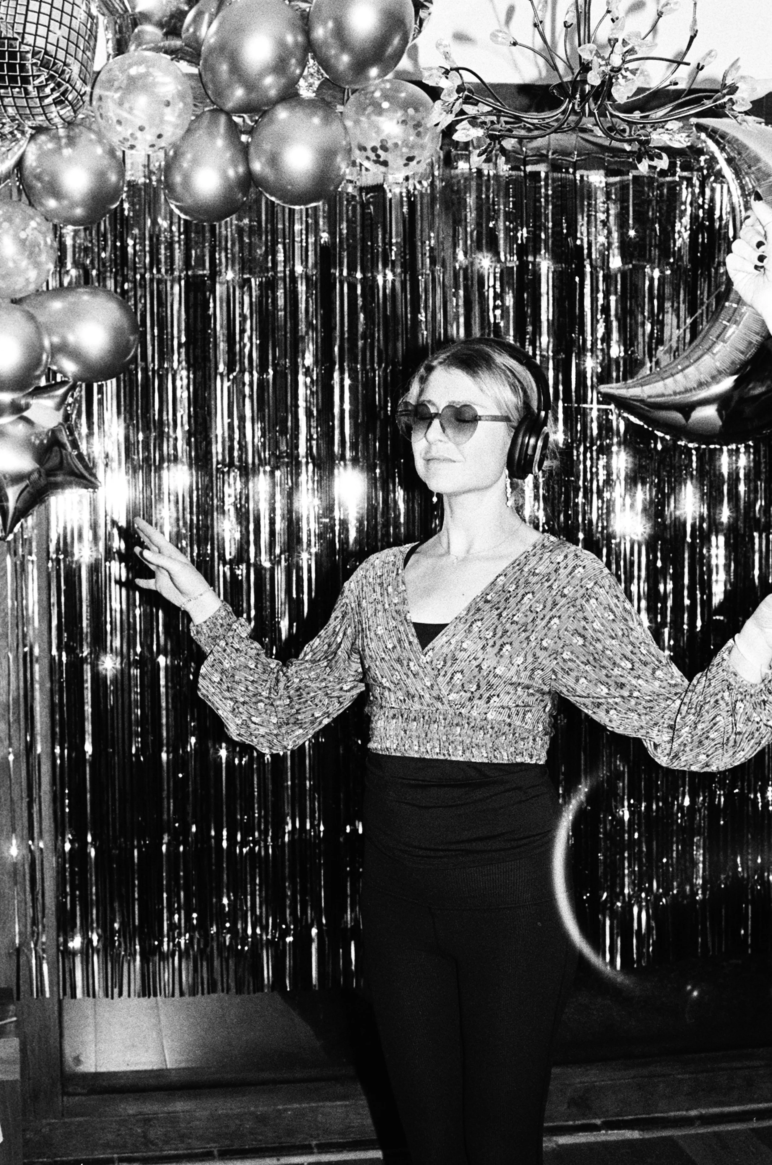 A woman wearing sunglasses and headphones dancing in front of a festive backdrop with balloons, metallic streamers, and decorative balloons, celebrating an event or party.