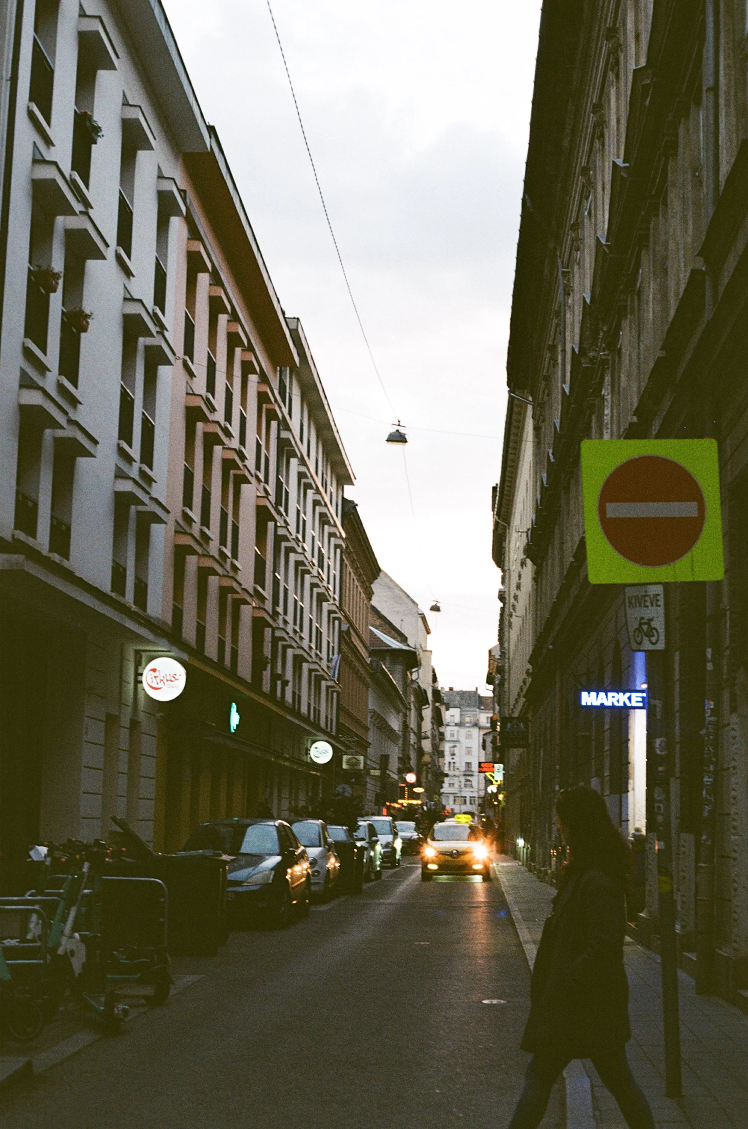 A street scene in a city during dusk with parked cars, illuminated shop signs, and a silhouette of a person walking.