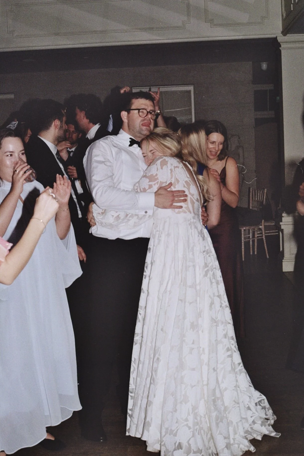 Guests dancing and celebrating at a wedding reception, with a man in glasses and a tuxedo hugging a woman in a white dress.