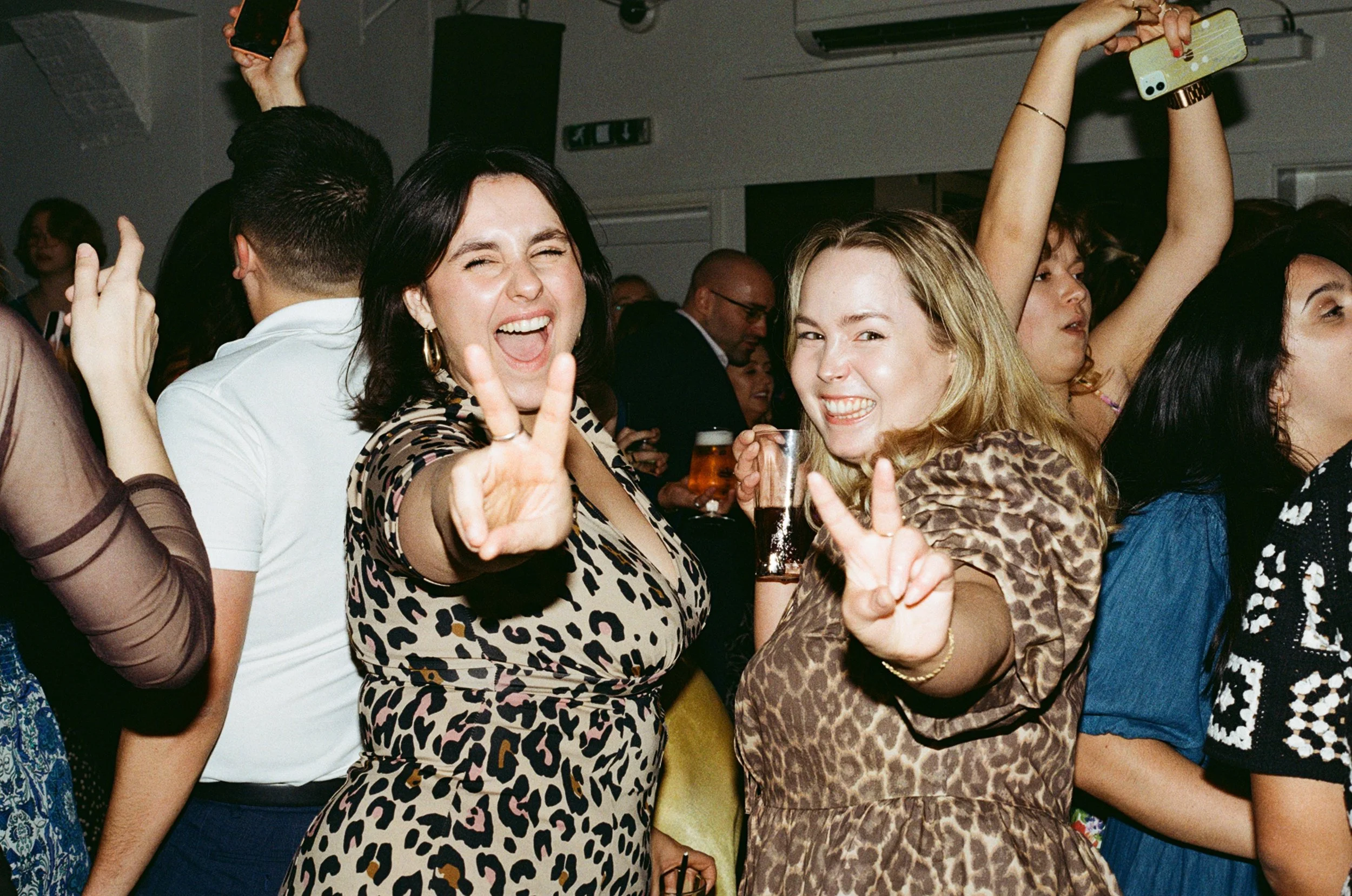 Two women in leopard print dresses posing and smiling at a party, making peace signs with their hands, surrounded by other party guests.
