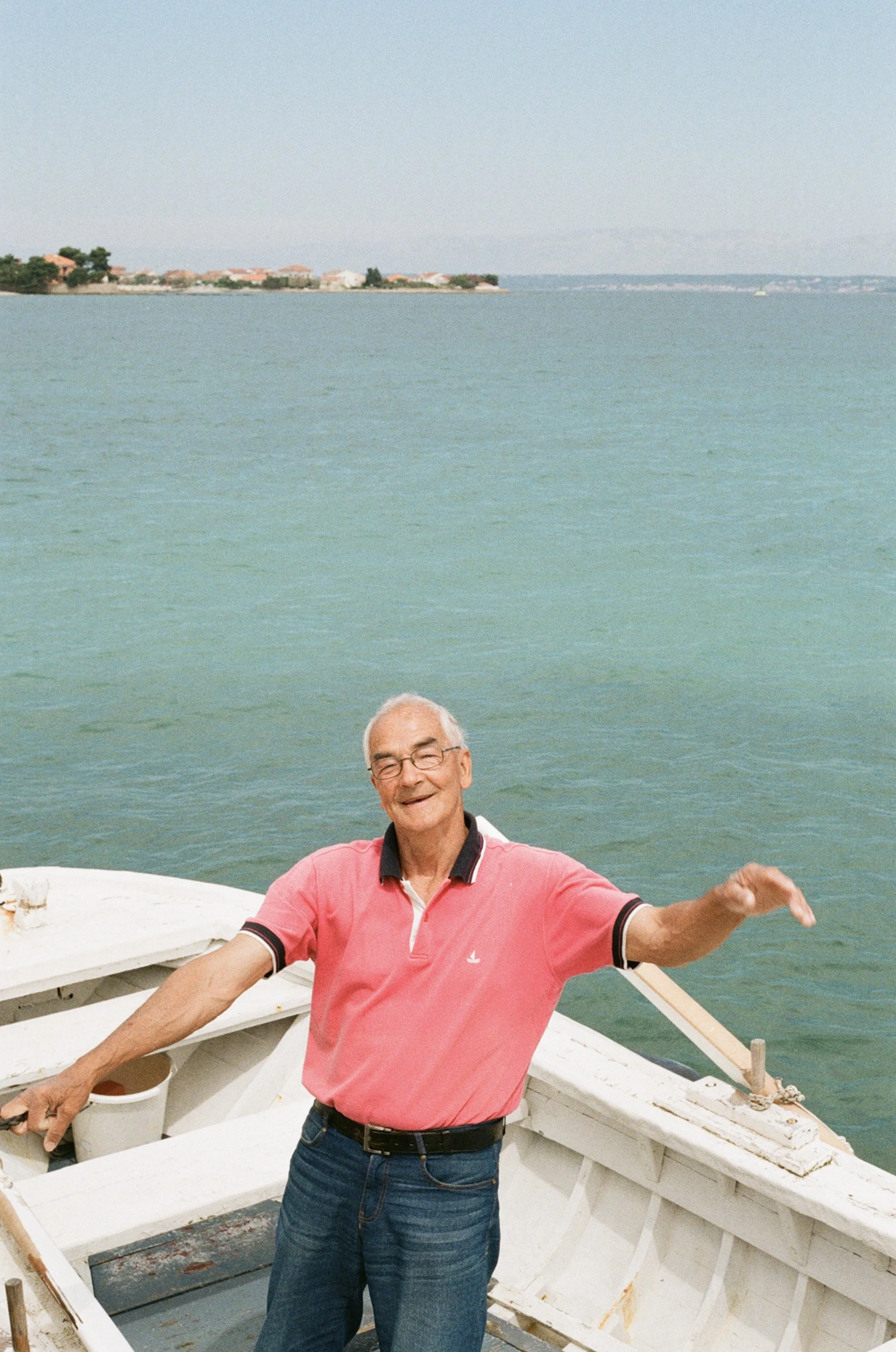 Older man on a boat smiling, wearing glasses and a pink polo shirt, with water and a distant shoreline in the background.