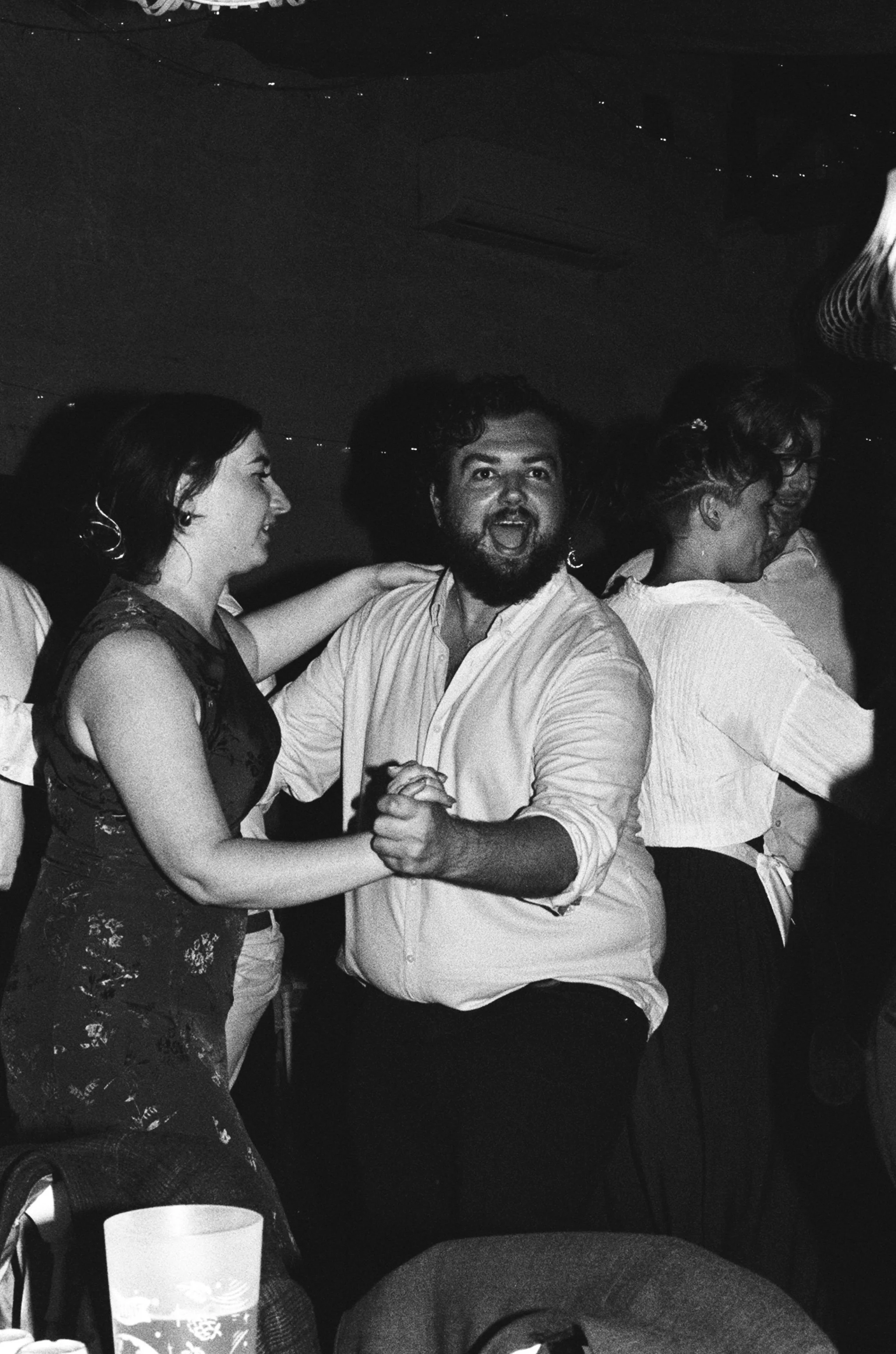 People dancing at a social gathering, with one man and woman in the foreground holding hands and smiling, others dancing in the background.