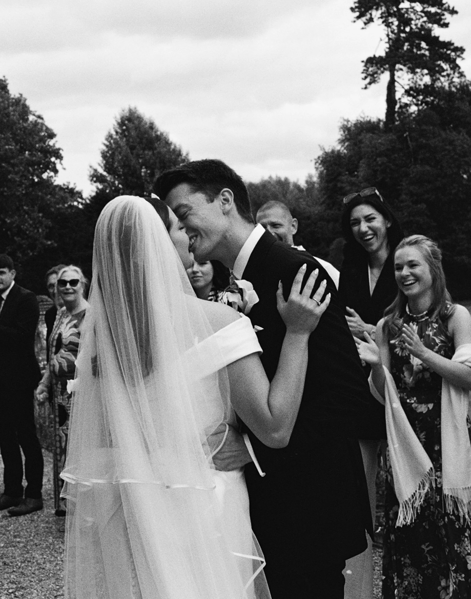A black-and-white photo of a bride and groom sharing a dance at their outdoor wedding. Several guests are around them, smiling and clapping, with trees and cloudy sky in the background.