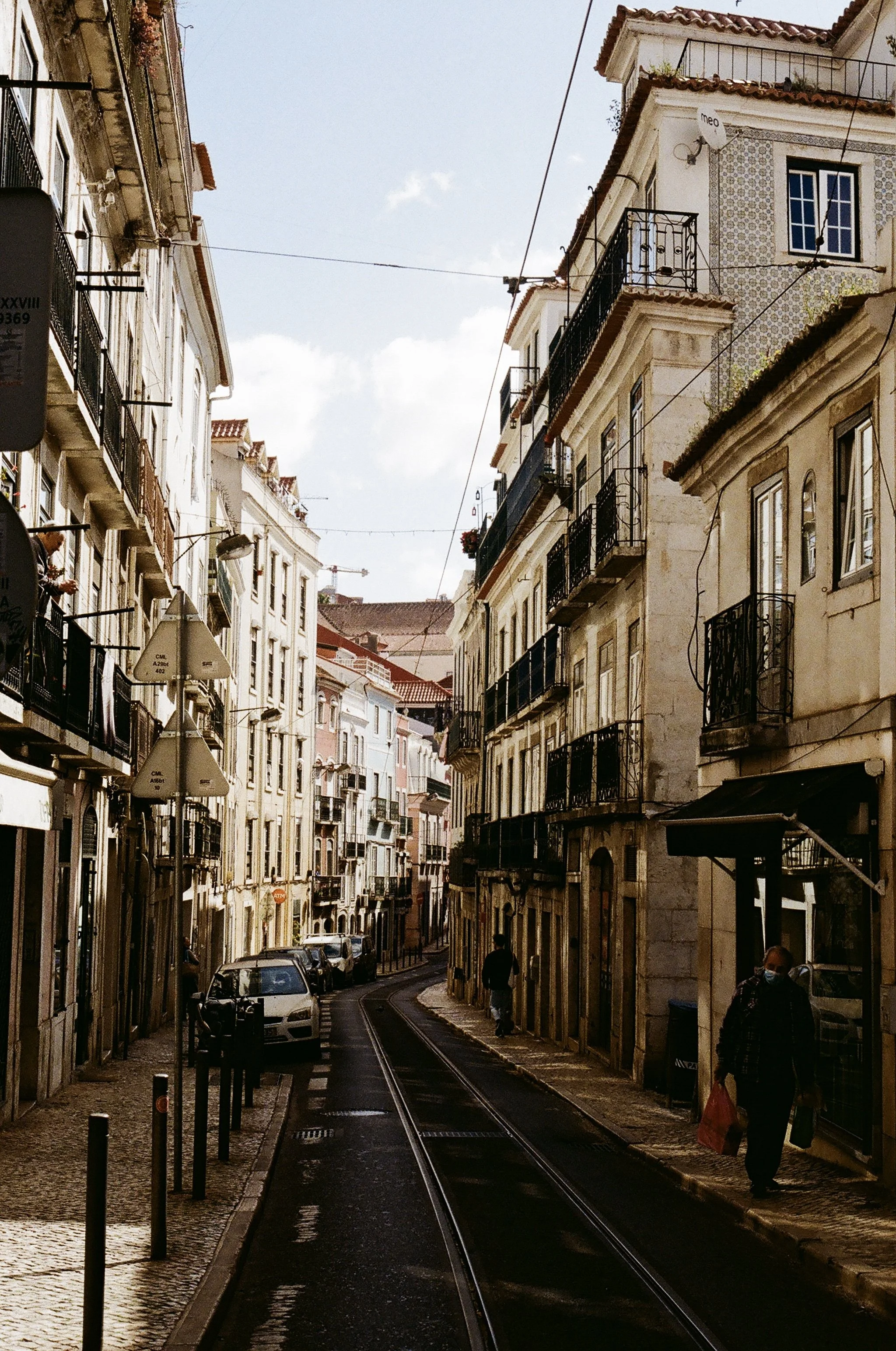 A narrow city street with tram tracks, parked cars, and multiple-story buildings with balconies, some with laundry hanging, under a partly cloudy sky.