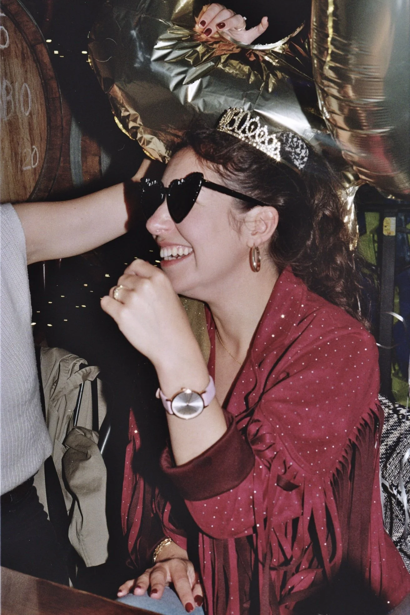 A woman party celebrating, wearing a 'birthday' tiara and heart-shaped sunglasses, smiling with a woman touching her face, set against birthday balloons and decorations.