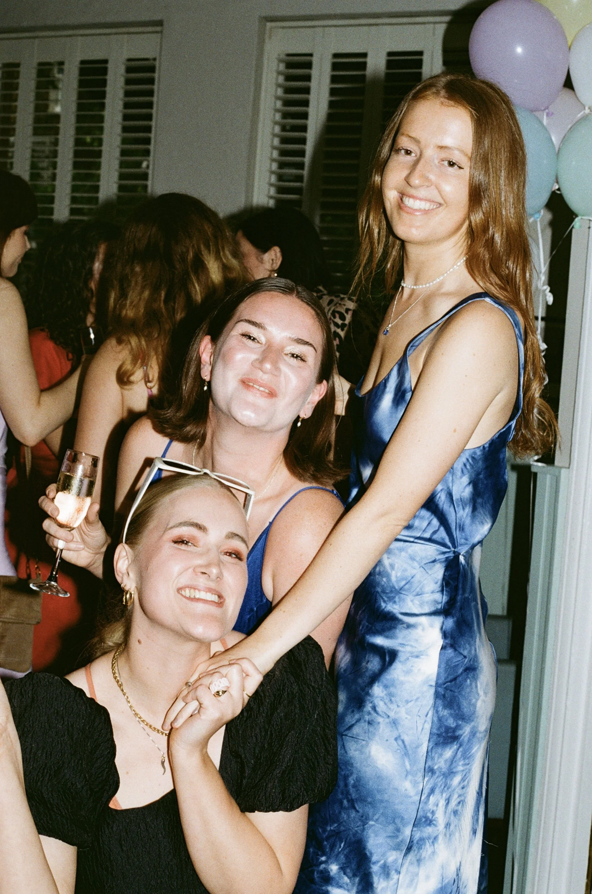 Three women smiling and posing closely together at a celebration, with balloons in the background and other people visible behind them.