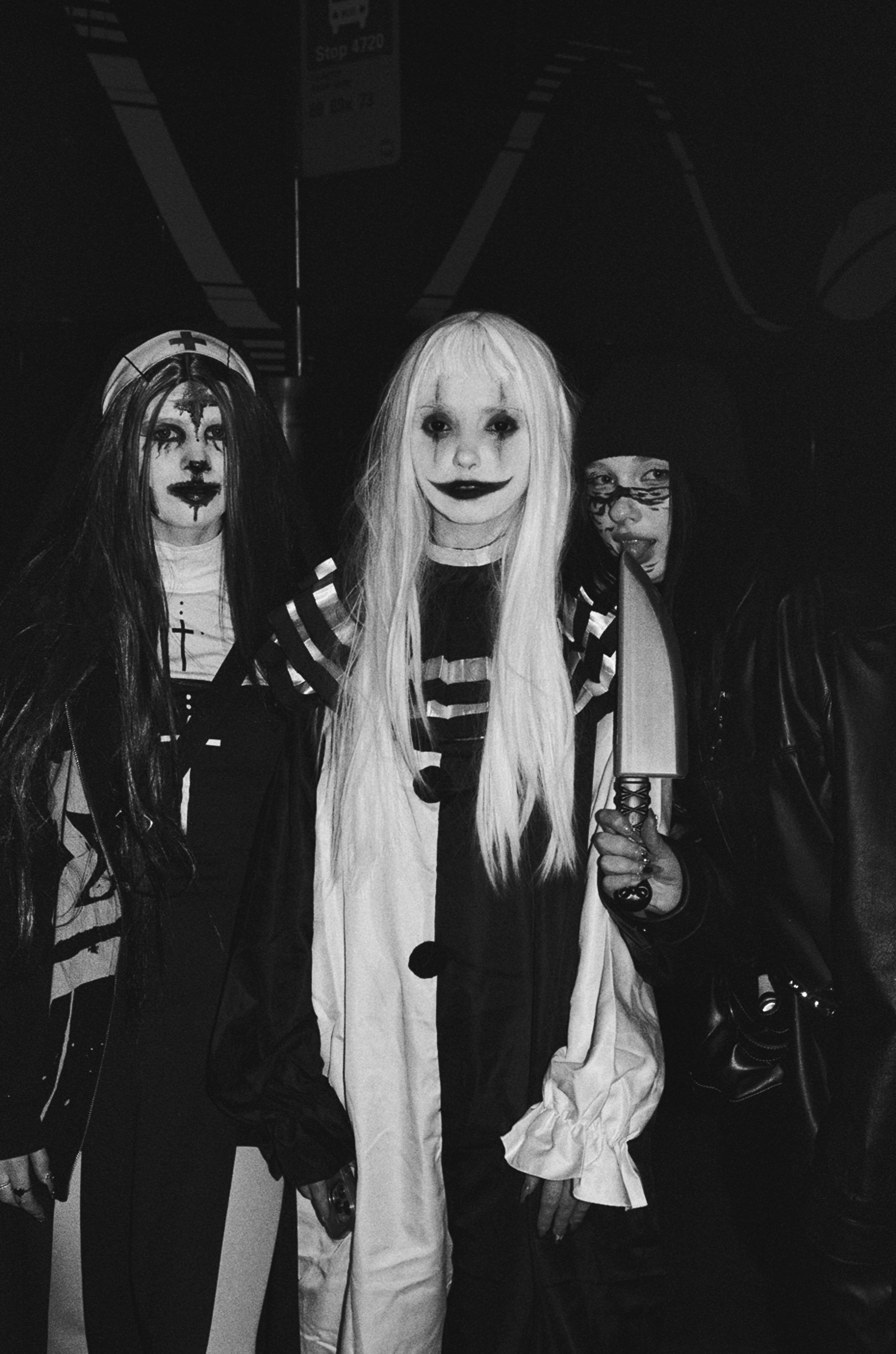 Three individuals in Halloween costumes. The person on the left dressed as a nun with a face painted to resemble a clown or jester. The person in the middle dressed as a clown with long light hair, face makeup, and a striped costume. The person on th