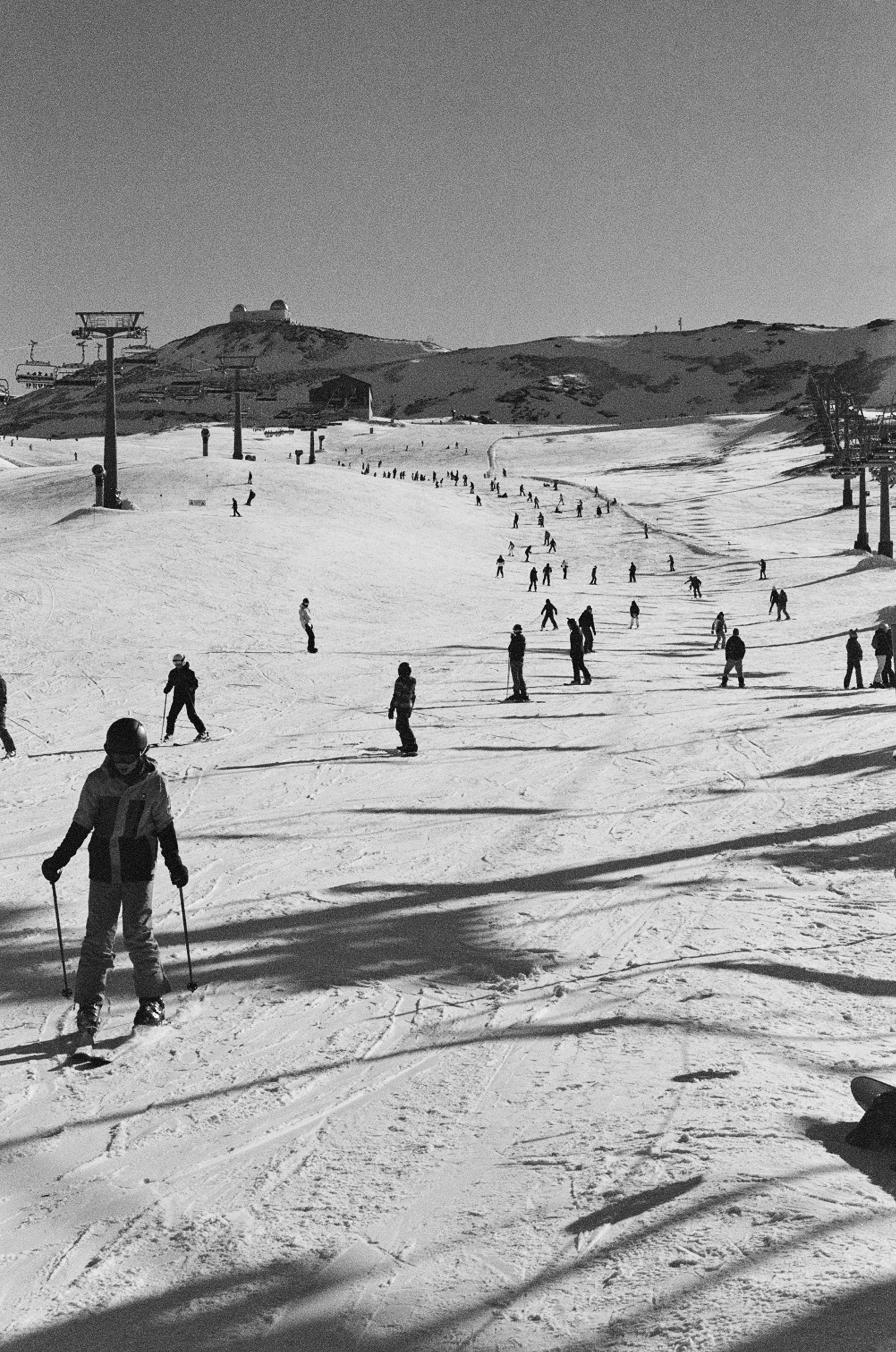 People skiing on a snowy mountain slope with ski lifts and a building on the hill in the background, in black and white.