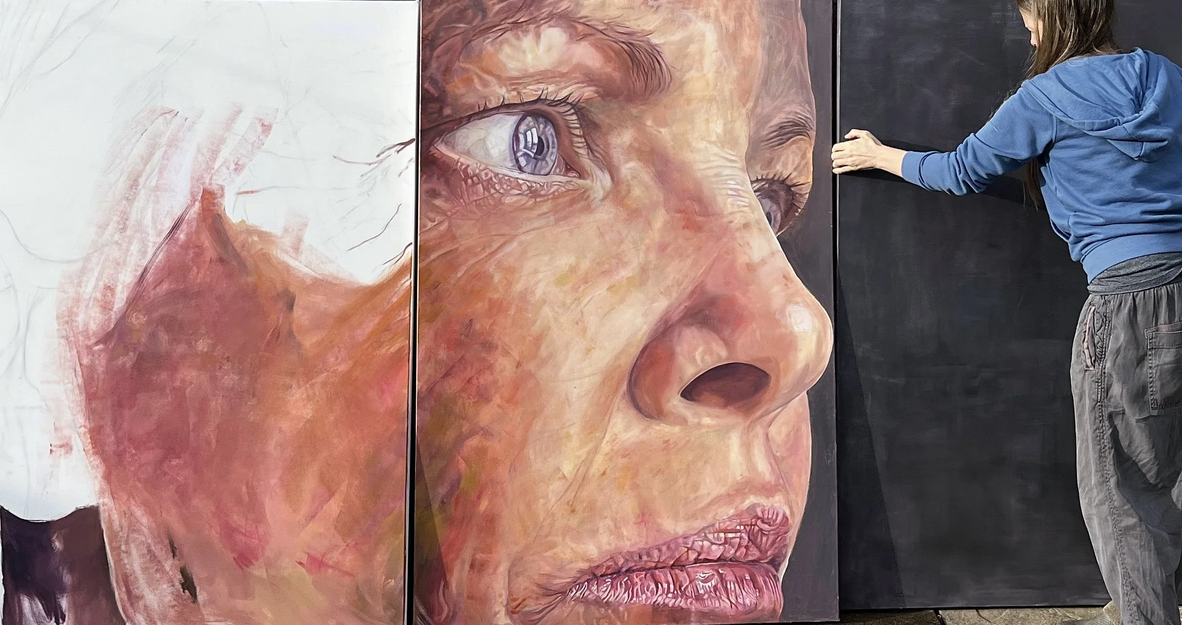 A woman in a blue hoodie and gray pants adjusting a large, three-panel painting of a realistic woman's face, focusing on the eye and nose area.