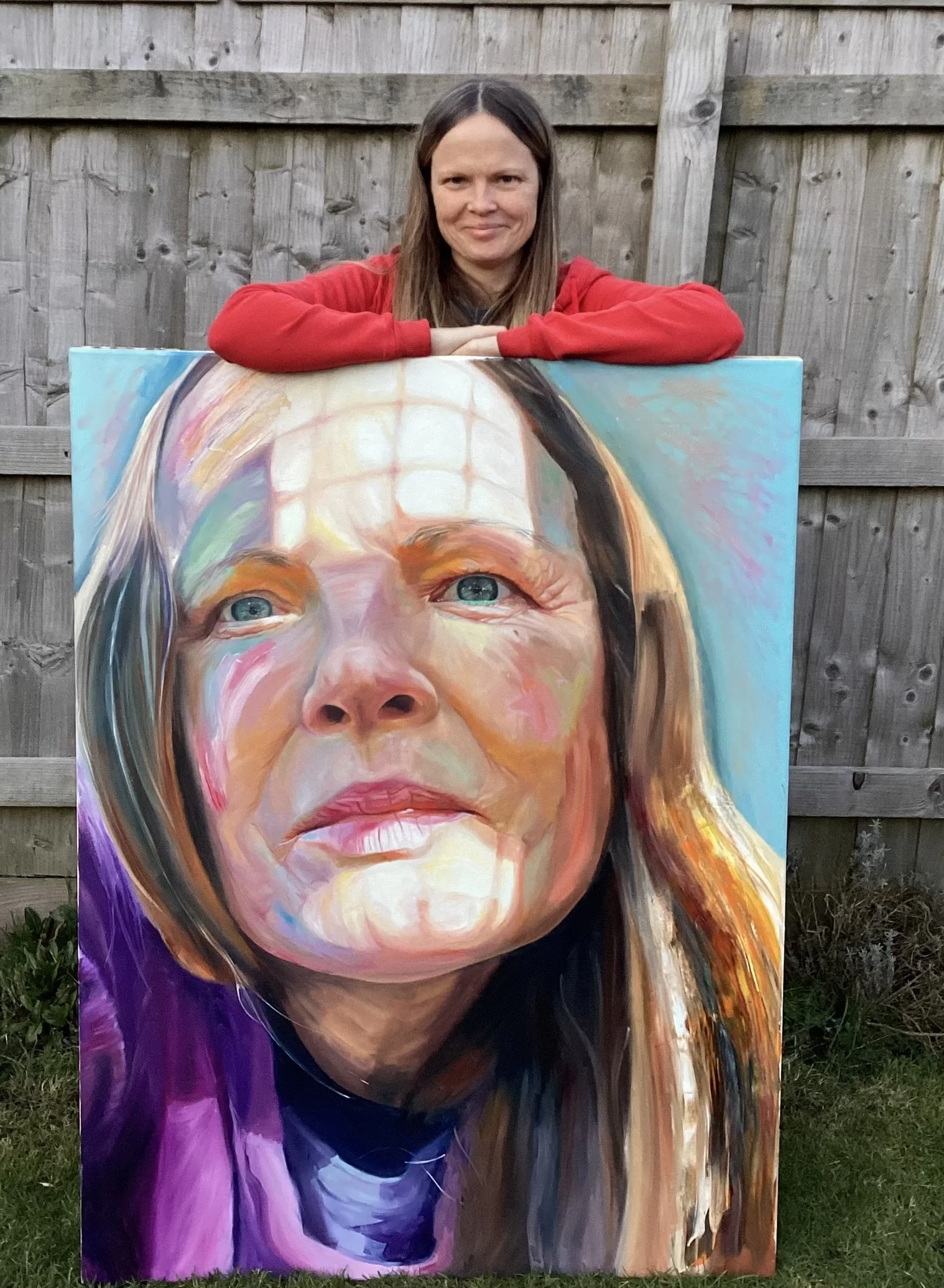 A woman in a red shirt is leaning on a large colorful portrait painting of a woman's face, outdoors in front of a wooden fence.