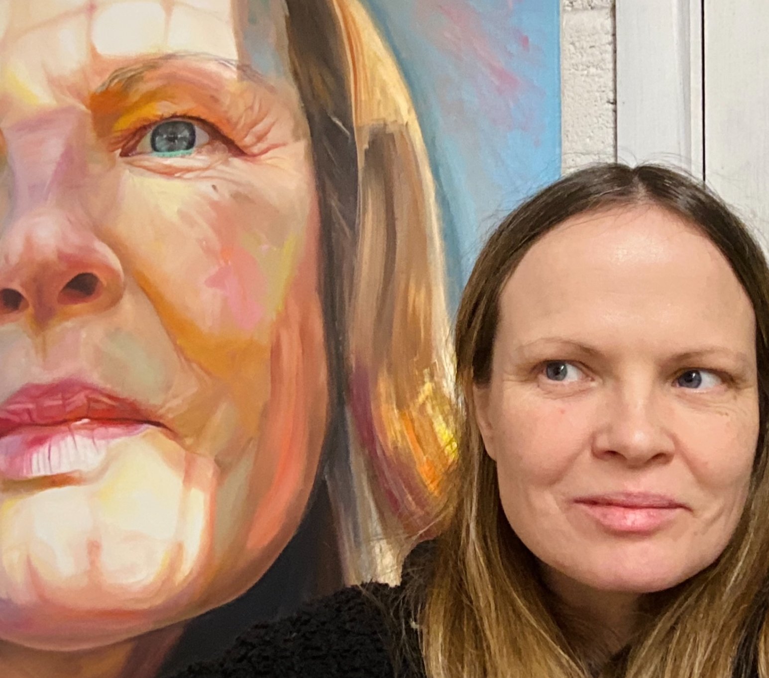 A woman with brown hair and blue eyes standing next to a colorful close-up portrait of a woman's face.