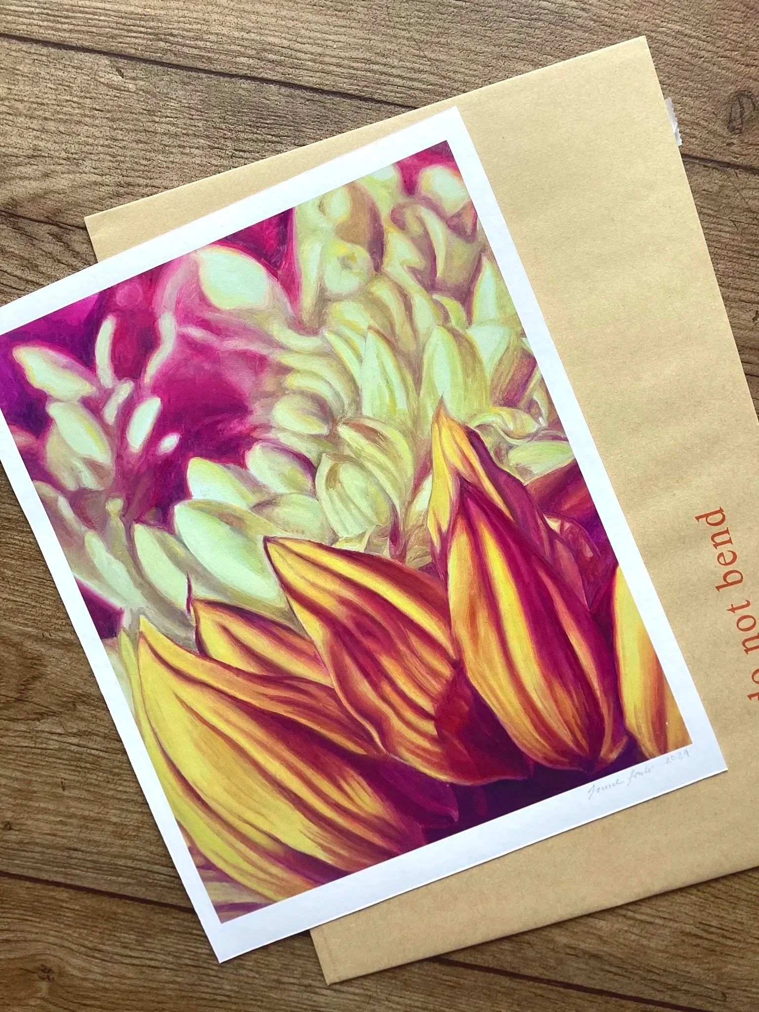 Colorful watercolor painting of a yellow and purple flower on white paper, placed on a brown paper envelope on a wooden surface.