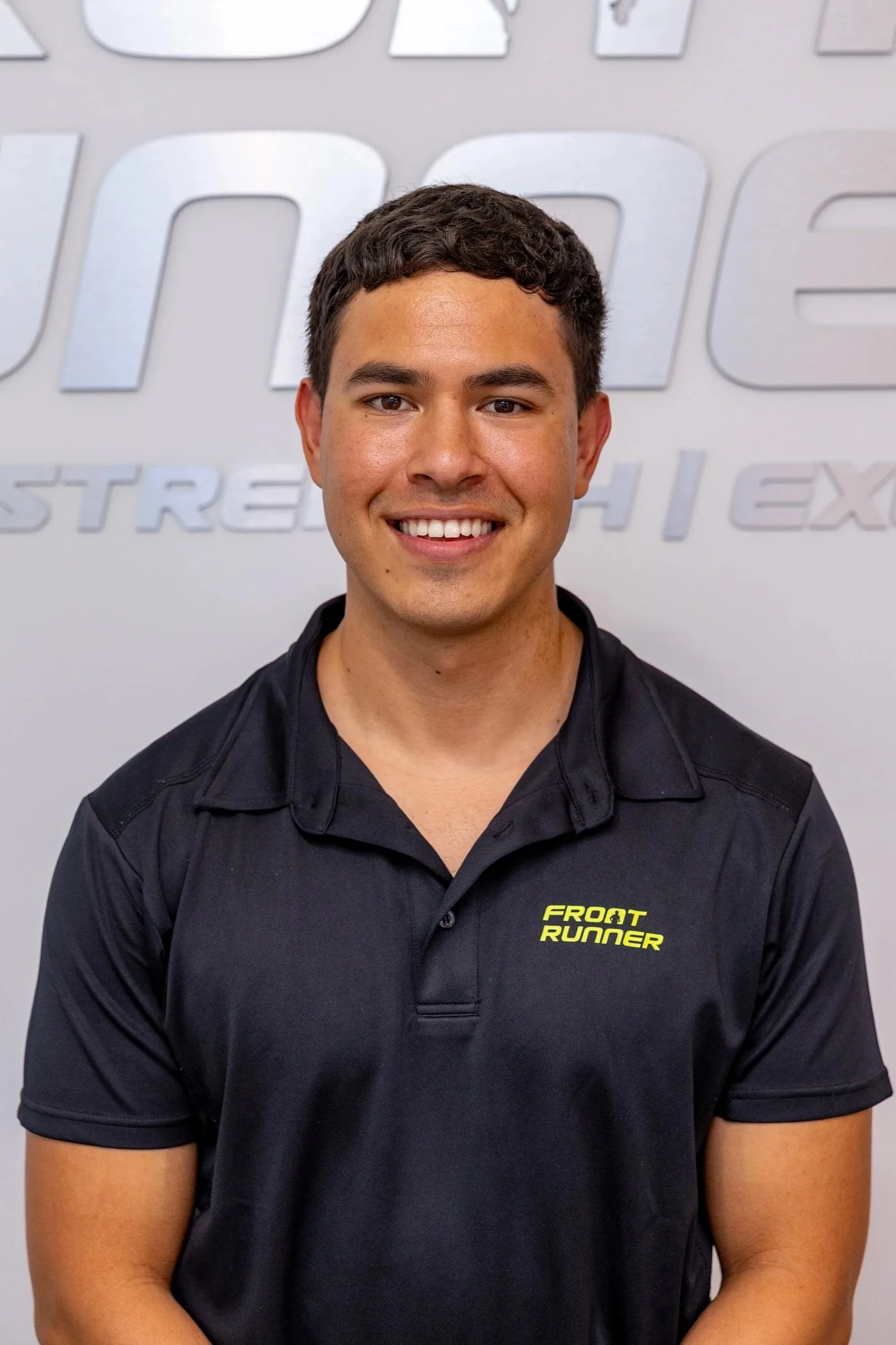 smiling young man in black polo shirt with yellow text that says 'Front Runner', standing in front of a wall with the logo 'OREGON TRAINING CENTER' in large silver letters.