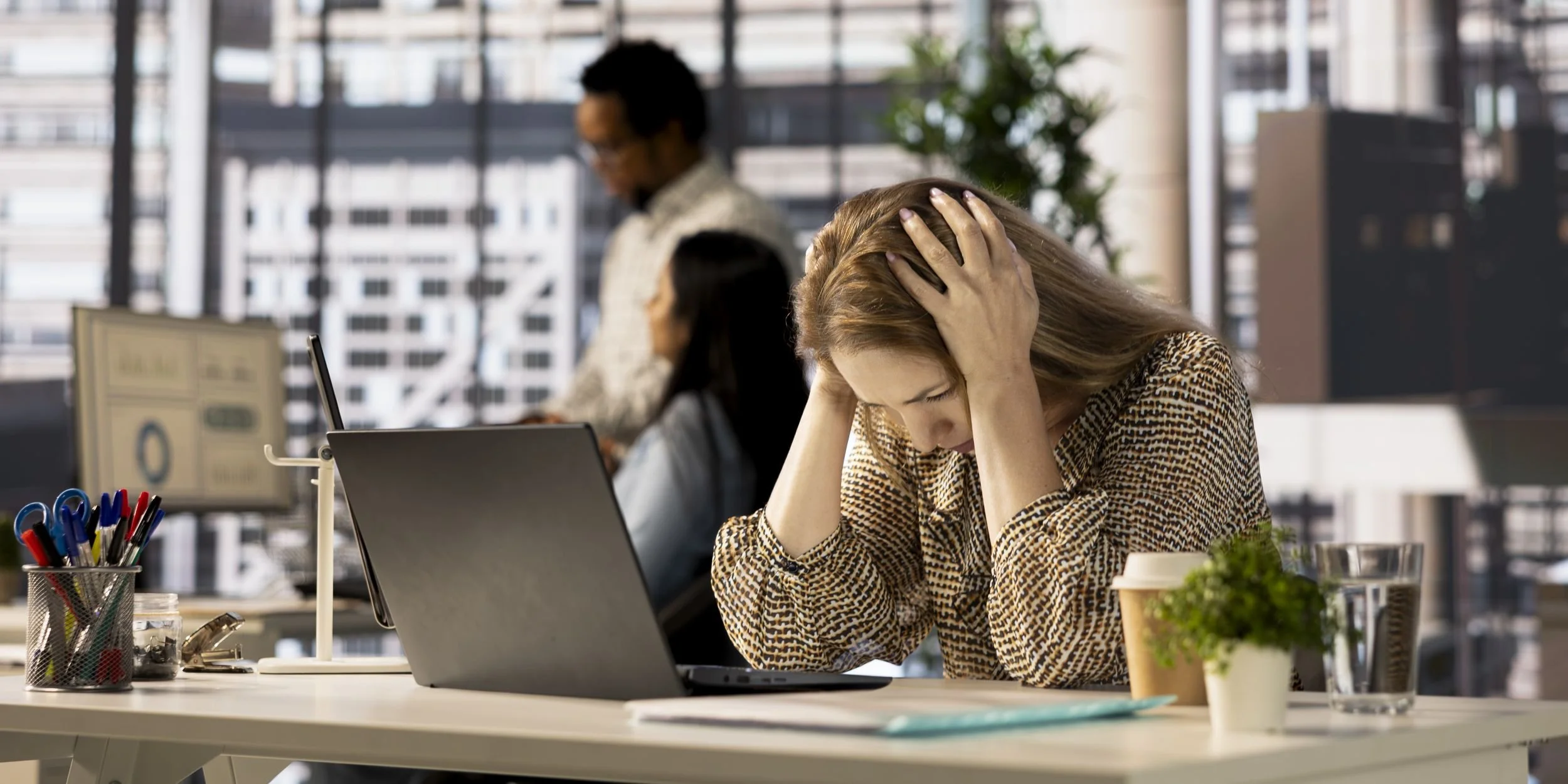 A stressed woman sitting at a desk in an office, holding her head with both hands, with a laptop, a coffee cup, pens, and papers on the desk, other colleagues working in the background.