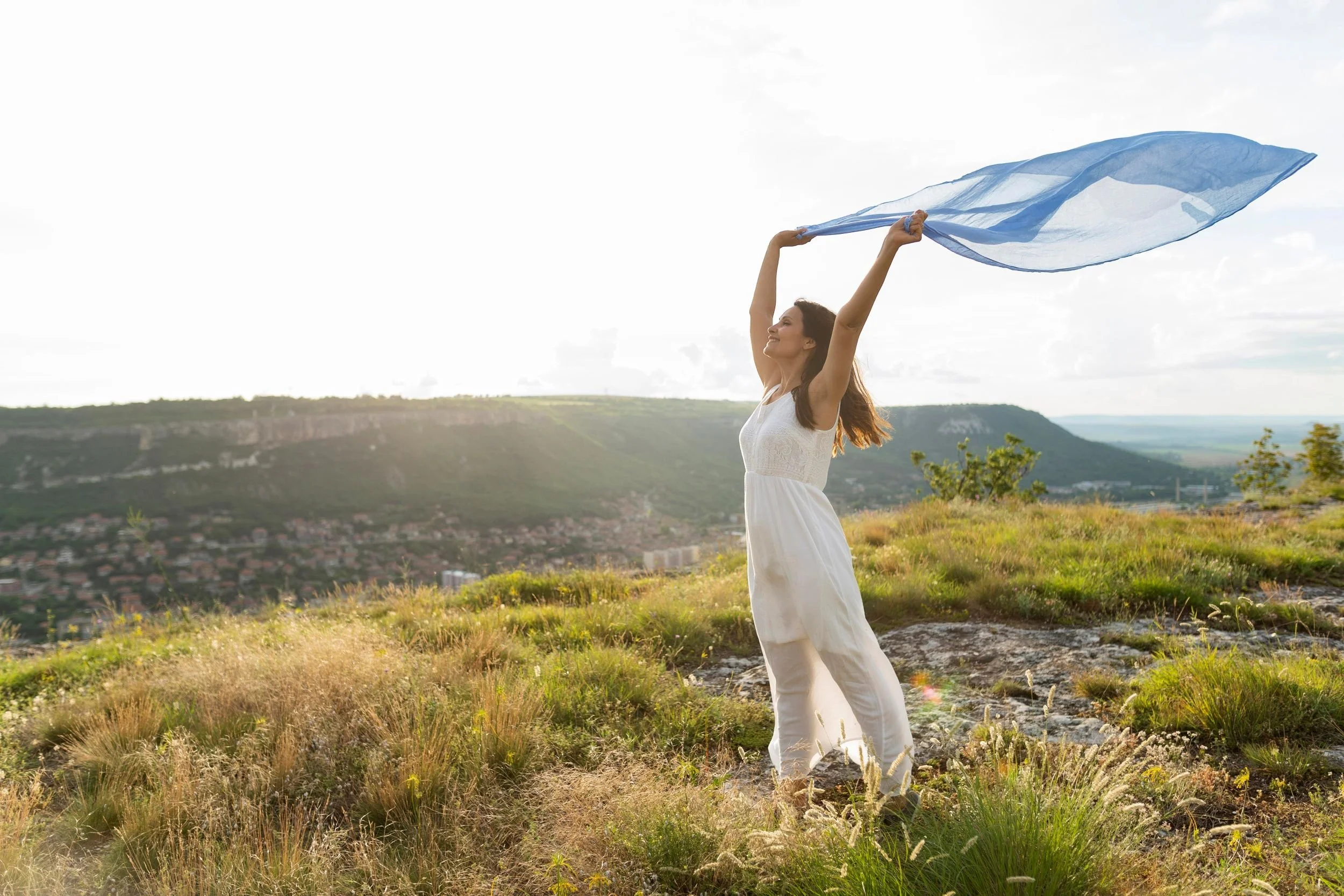 Woman standing on a grassy hill with her arms raised, holding a blue scarf in the wind, overlooking a landscape of hills and a small town at sunset.