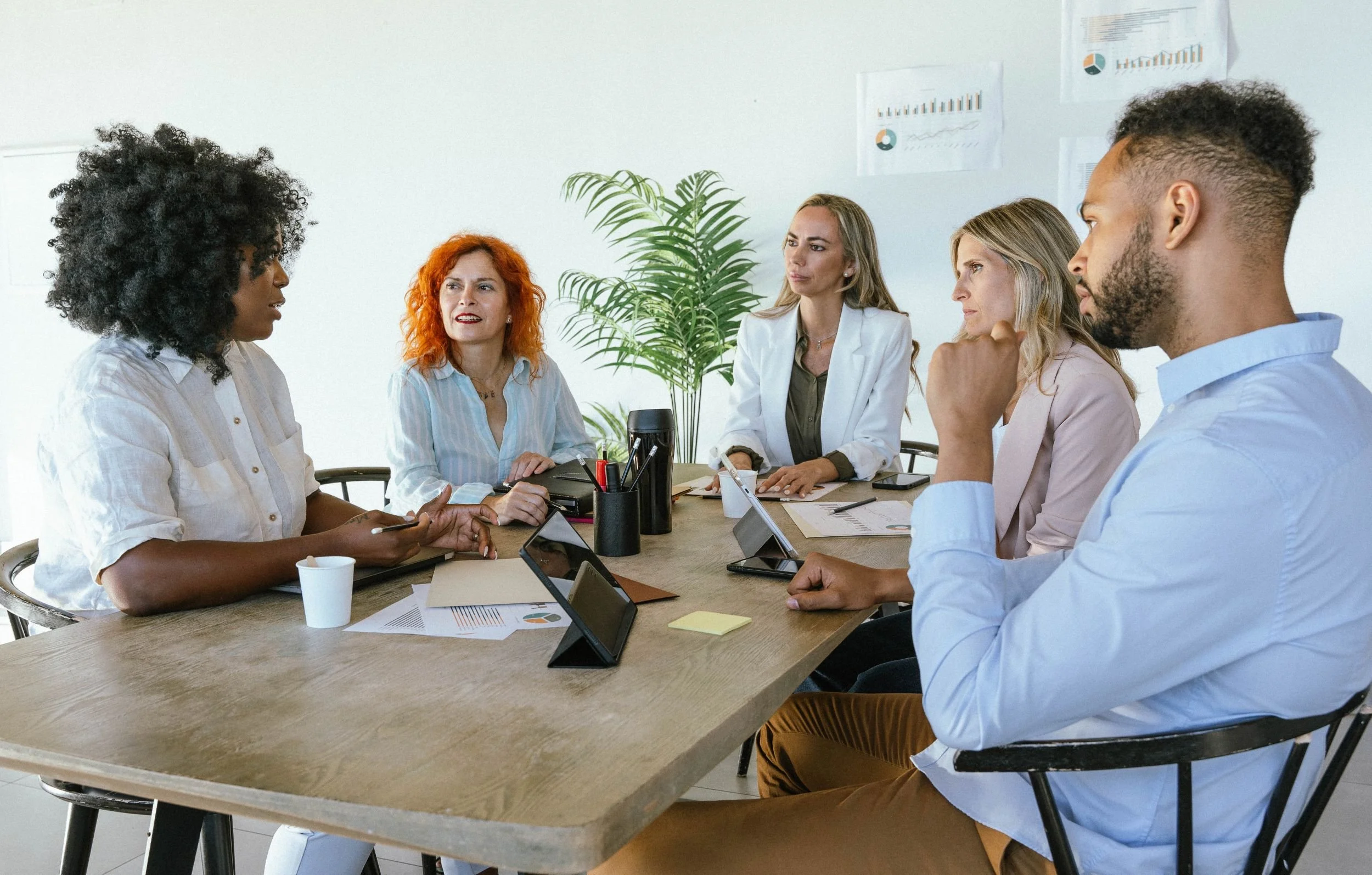 Six diverse people having a meeting around a wooden table in a bright office with charts on the wall and a potted plant.
