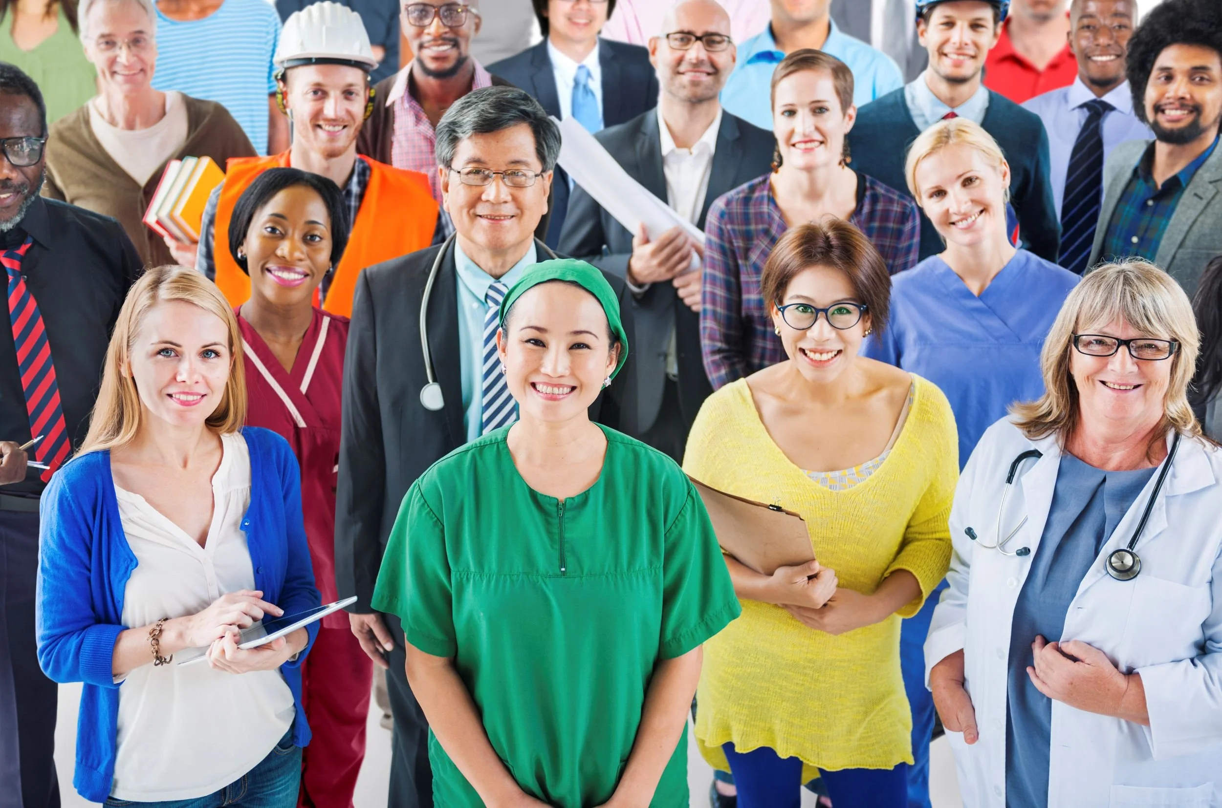 A diverse group of medical professionals and staff in colorful scrubs, lab coats, and business attire, smiling and gathered together in a hospital or healthcare setting.