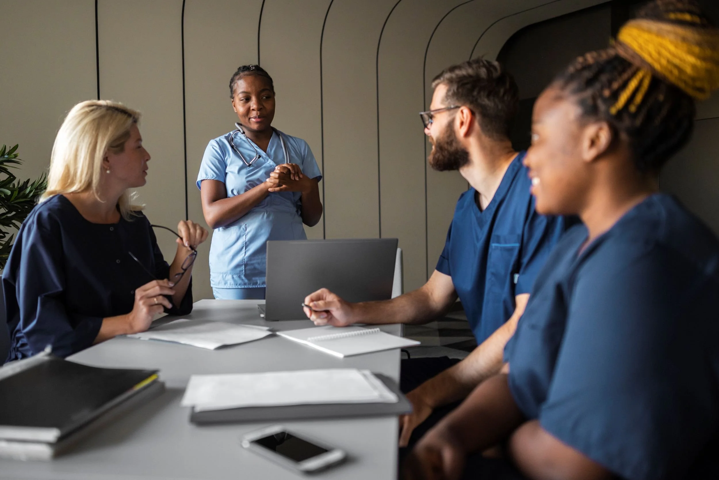 A group of nurses in a medical meeting, with a woman nurse in blue scrubs standing and speaking to three seated colleagues in blue scrubs around a table, with a laptop and notebooks.