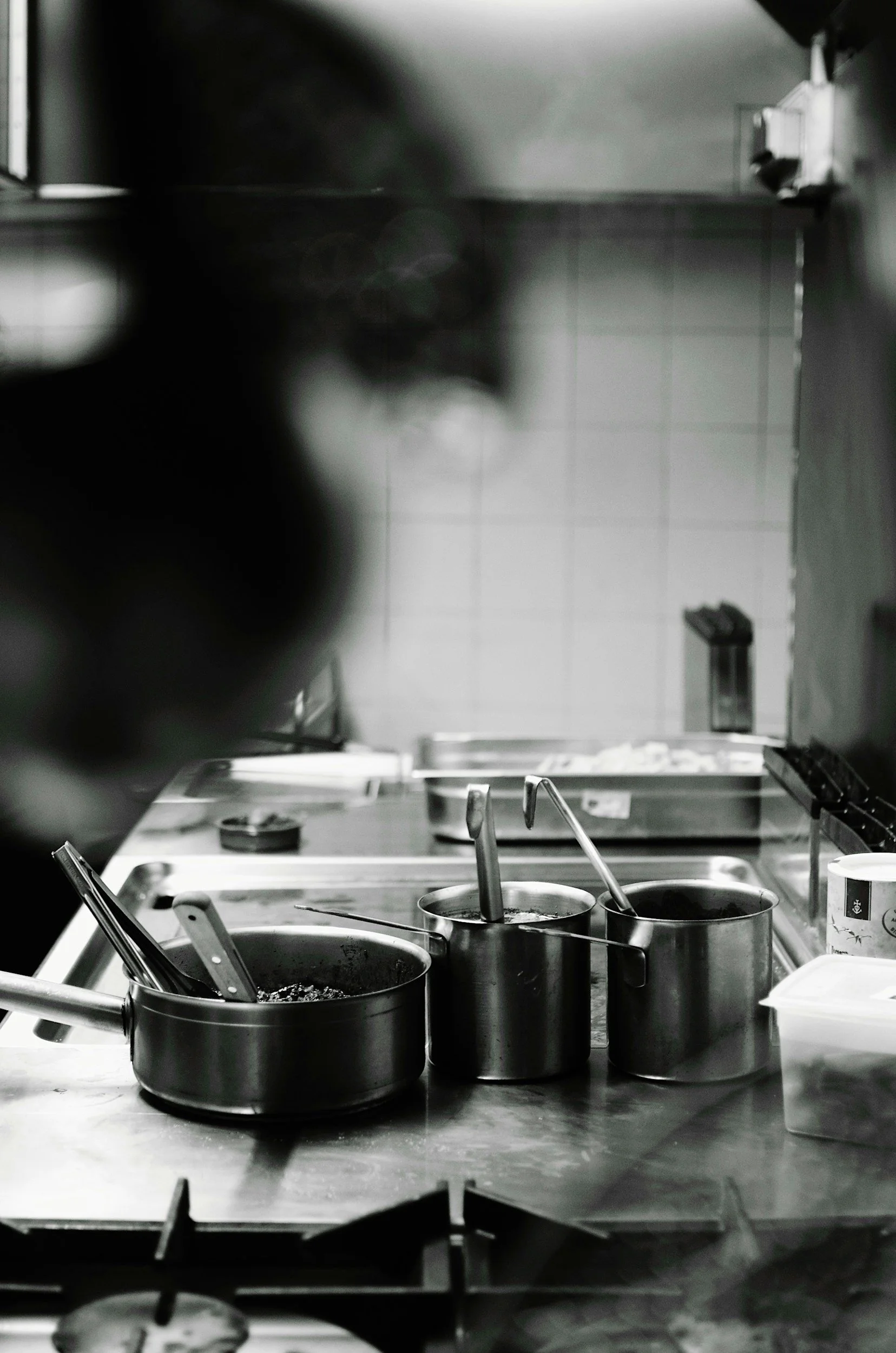 Black and white photo of a commercial kitchen counter with saucepans and utensils, blurred person in the foreground, tiled wall in the background.