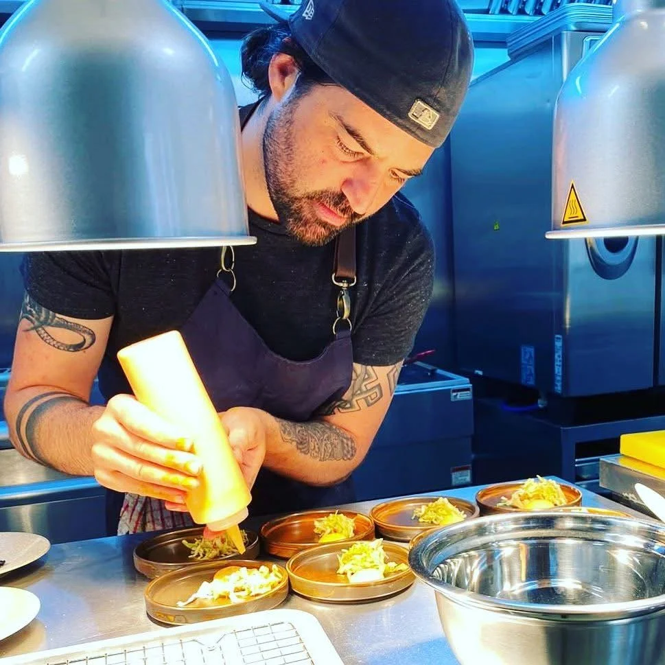 A man wearing a black cap and dark t-shirt is preparing food in a commercial kitchen, using a squeeze bottle to add sauce to dishes on a countertop.