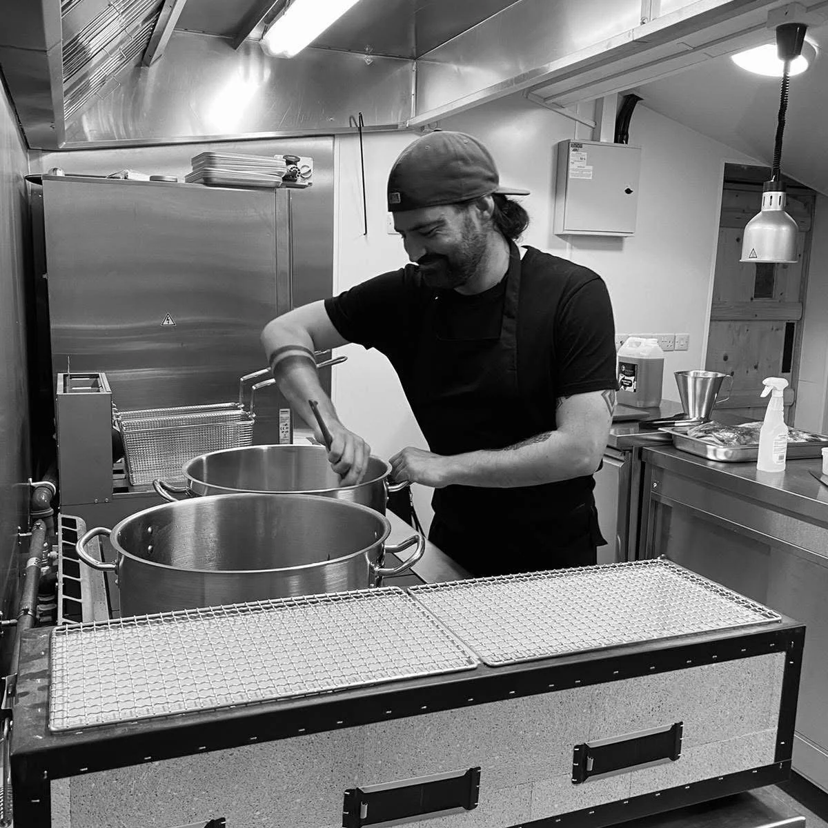 A man wearing a cap and apron cooking or preparing food in a commercial kitchen, stirring a large pot on a stove.