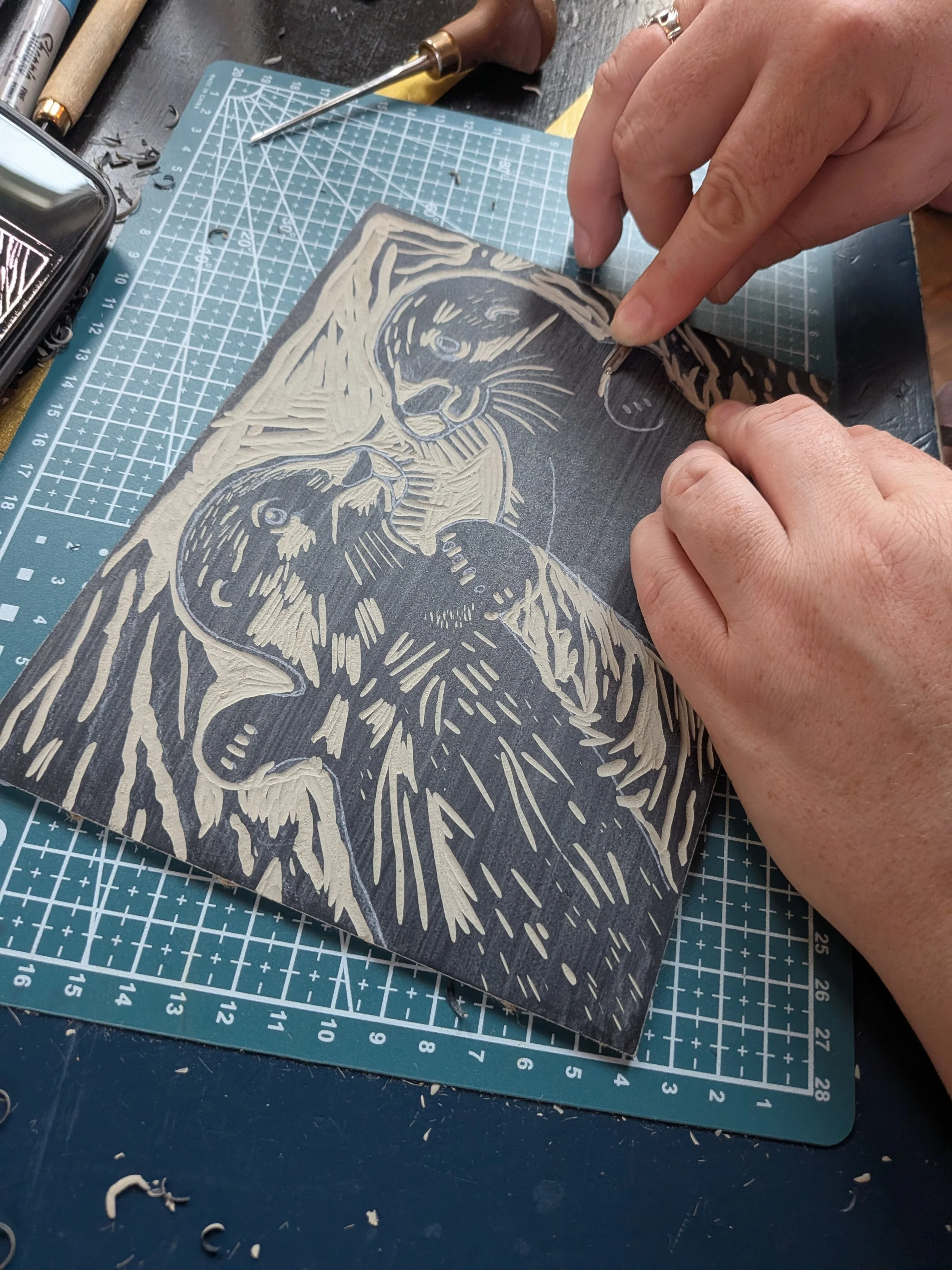 Hands carving a detailed bird design into a linoleum block with a gouge tool on a gridded cutting mat