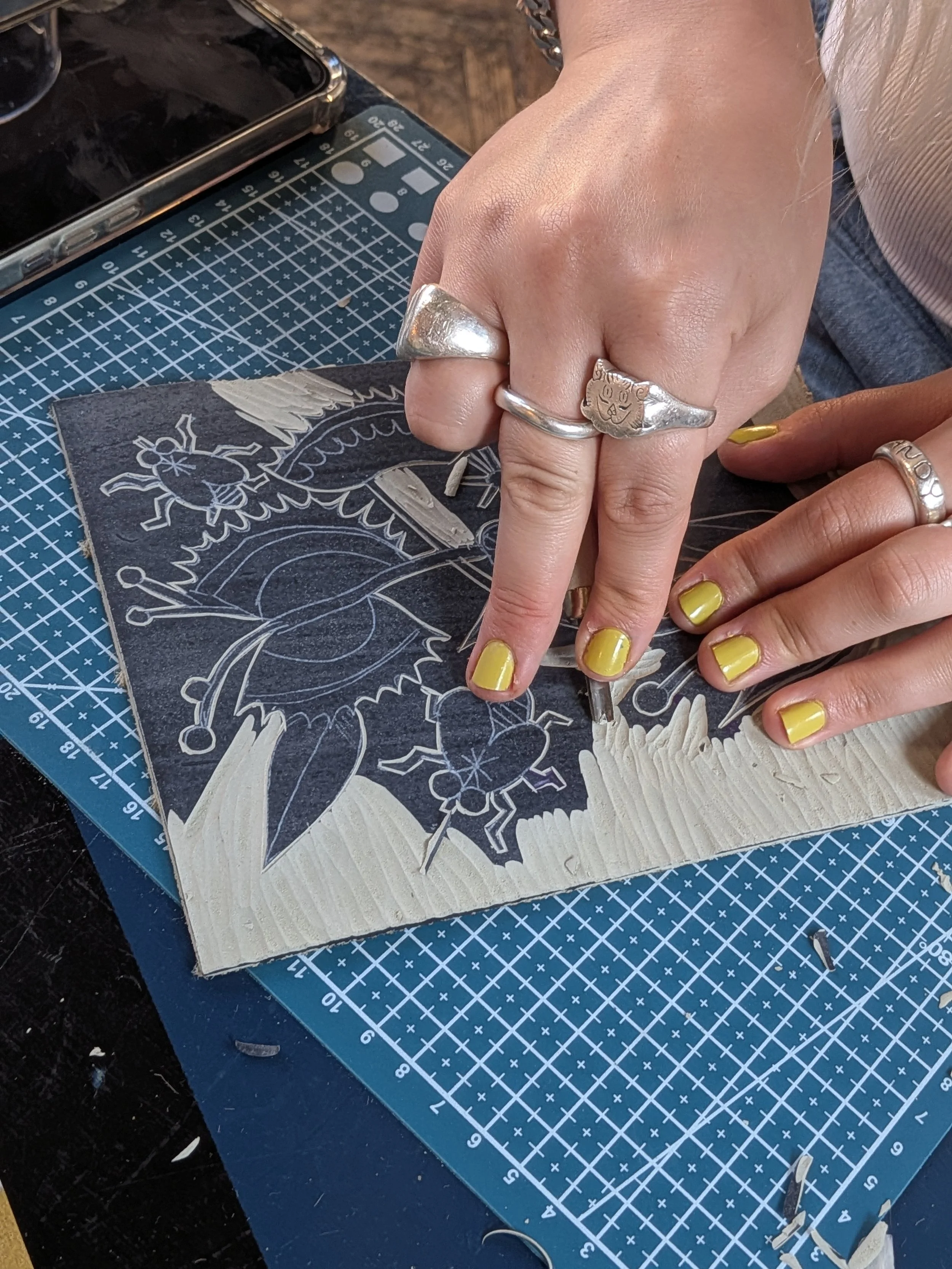 Hands with yellow-painted nails use a craft knife to cut white textured material on a patterned cutting mat.