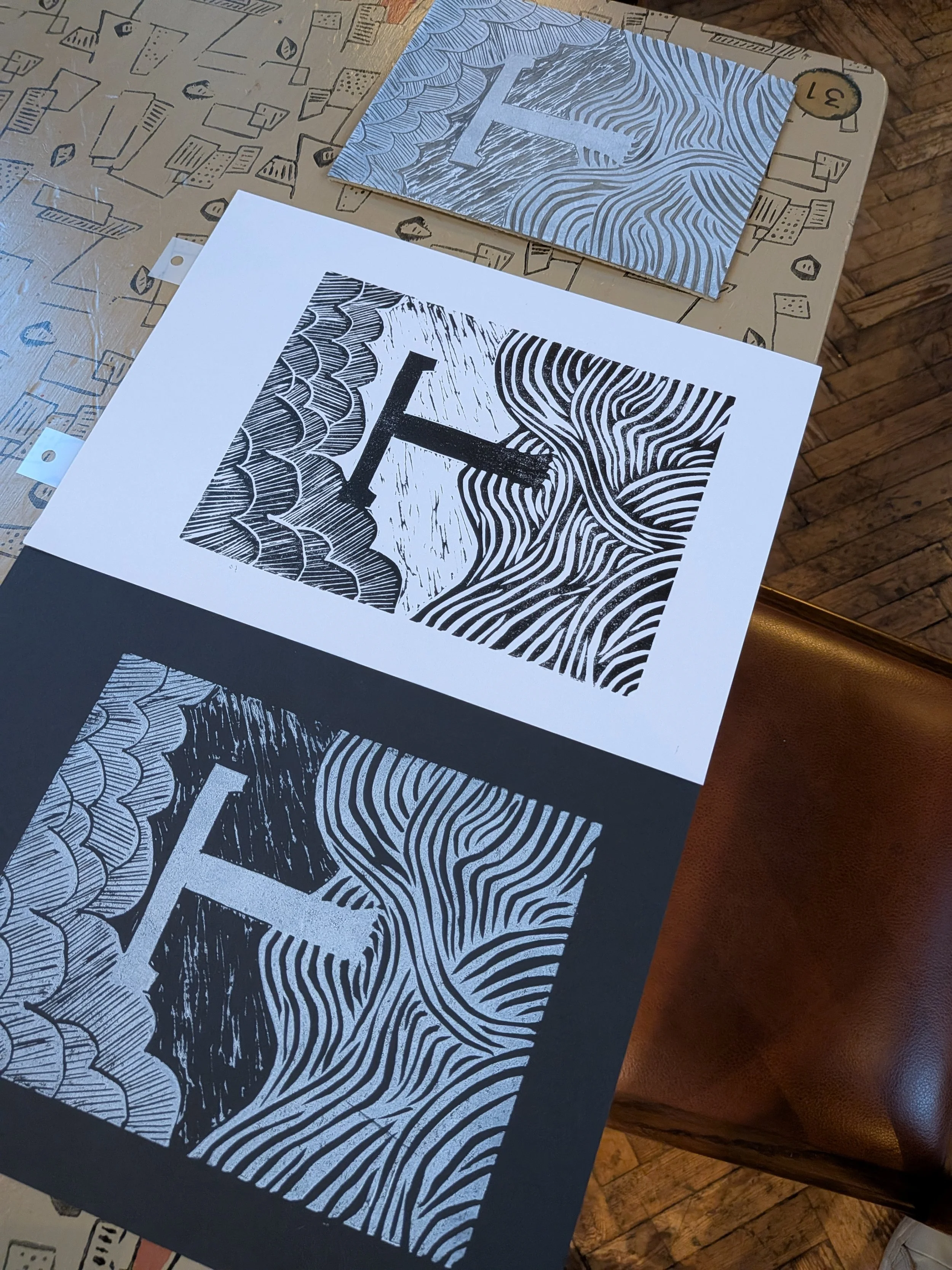Abstract black-and-white block print artwork with bold geometric shapes, wavy lines, and scalloped patterns displayed on a patterned tabletop.