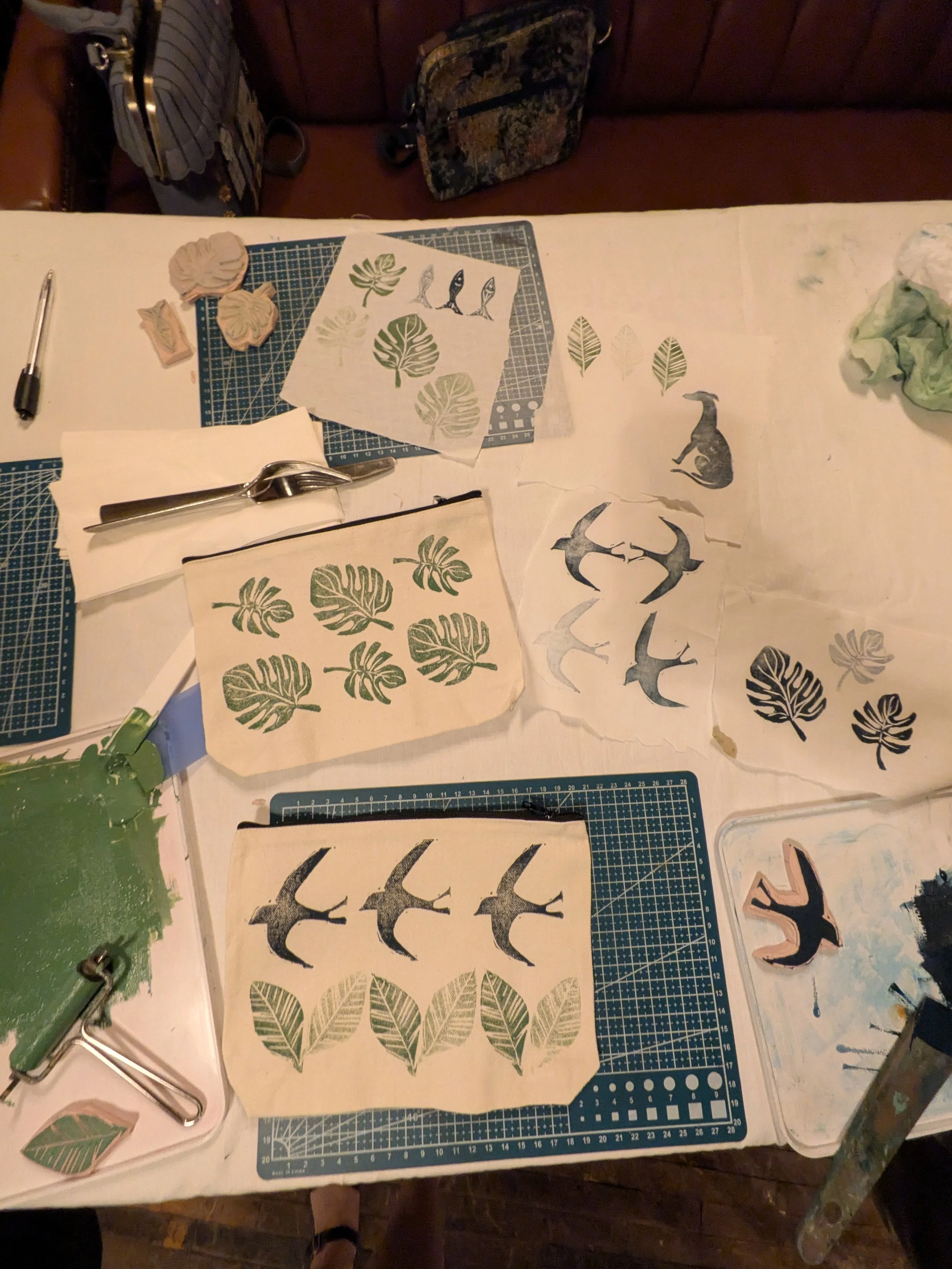 Artisan printmaking workspace with carved leaf and bird stamps, printed botanical designs, cutting mats, carving tools, and ink.