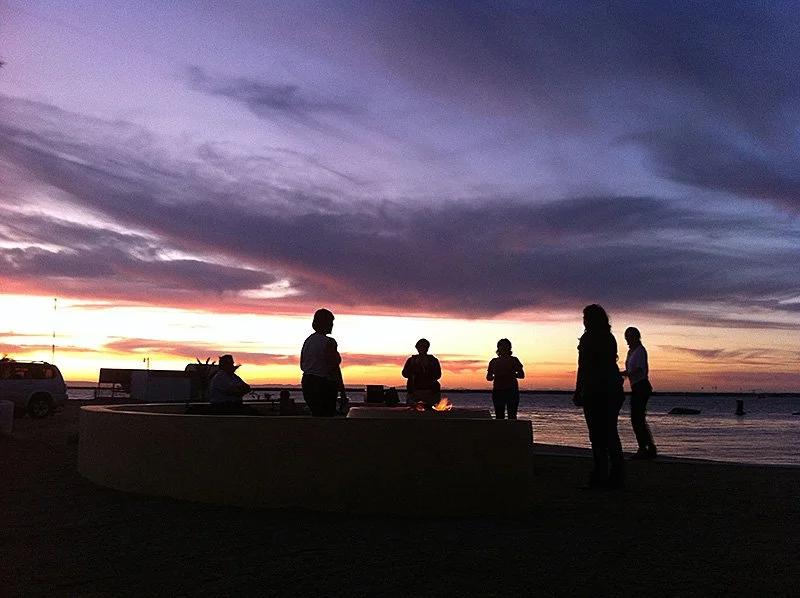 Silhouettes of six people standing at the edge of a large body of water, while pink, yellow, and gray clouds stretch across the sky dramatically at sunset or sunrise.