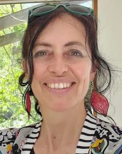 Marta Ceroni headshot. White presenting woman, smiling, wavy brown hair worn up, big red earrings, striped shirt.