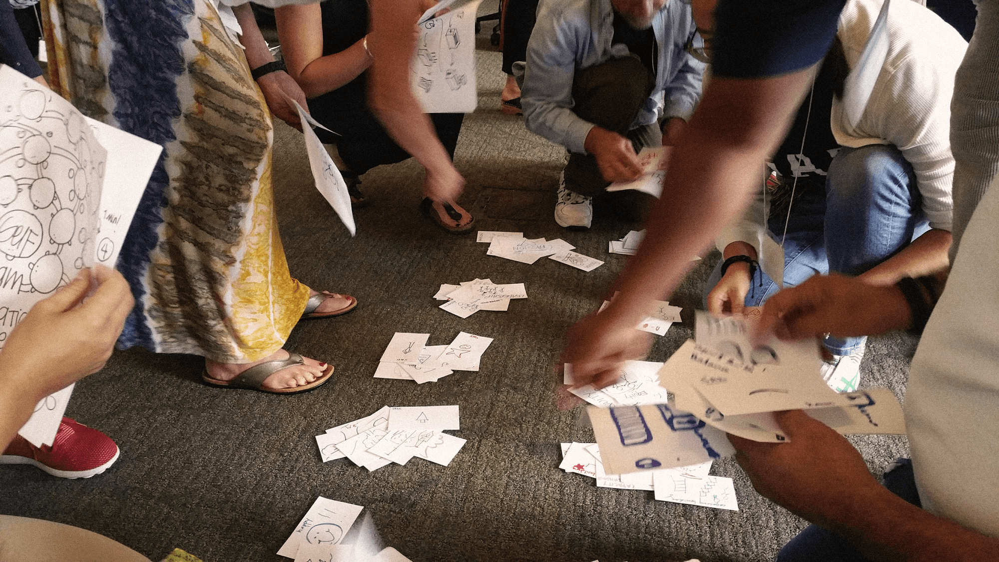 People gathered on the floor, exchanging and displaying various illustrated cards or papers, with some holding or reaching for the cards.