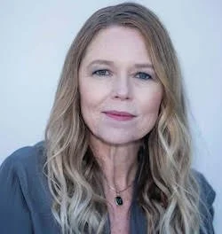 Carrie Norton headshot. White presenting woman, wavy blond hair worn down, grey top.