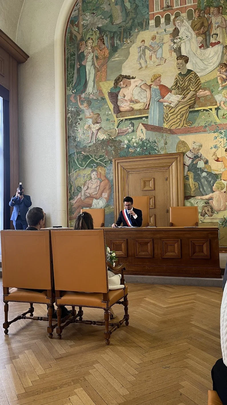 Une salle de conférence avec un homme en costume derrière un bureau en bois, portant une chaîne tricolore, et des personnes assises devant lui. Un grand tableau mural peint en arrière-plan représente plusieurs scènes de personnages dans un contexte classique ou mythologique.