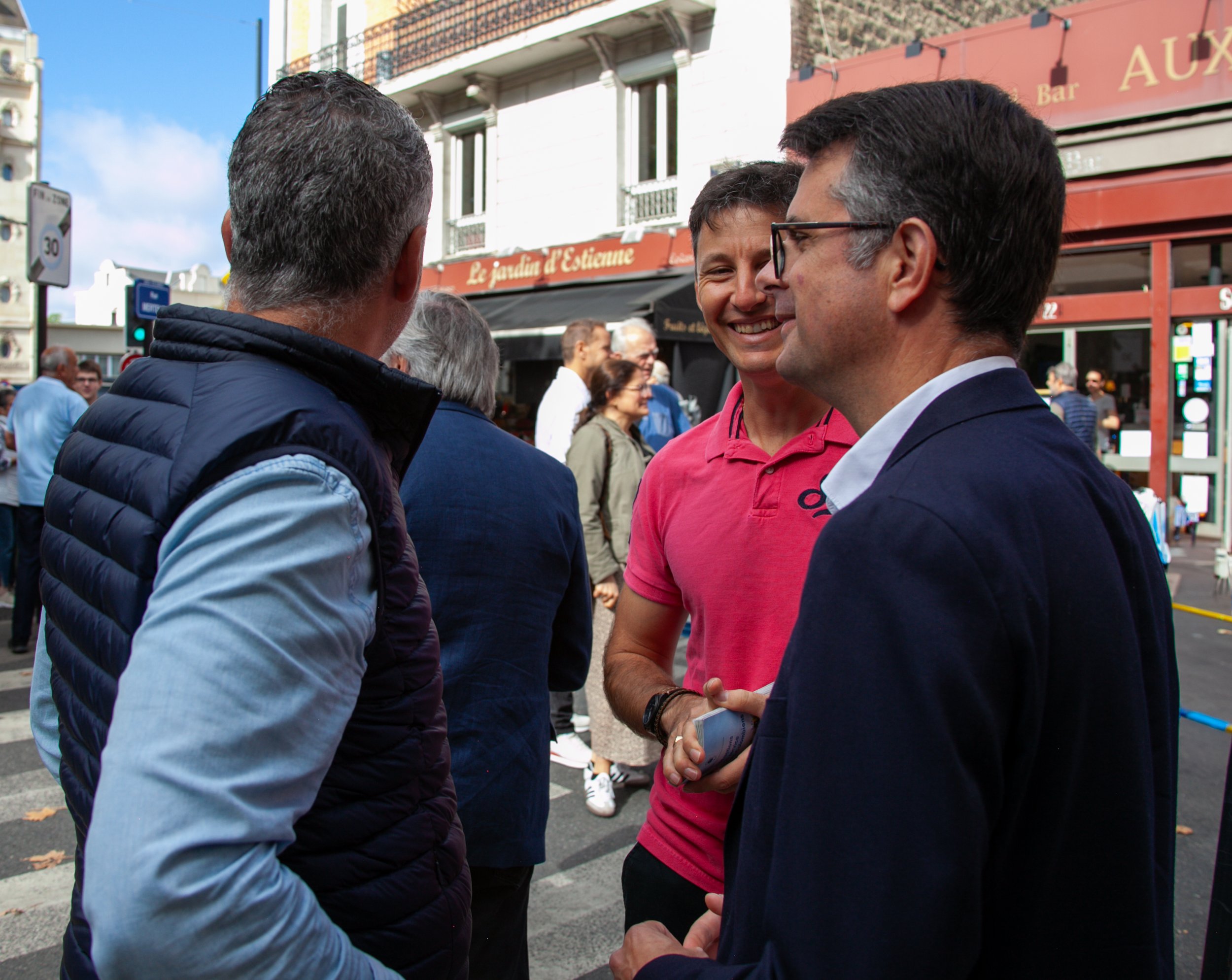 Trois hommes discutent et sourient dans une rue de Paris, avec des passants et des commerces en arrière-plan.