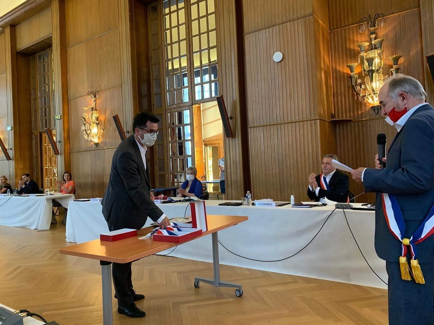 Une cérémonie officielle où une personne est en train de voter dans une enveloppe dans une pièce décorée en bois avec plusieurs personnes assises à une longue table. Deux hommes en costume avec bandeaux tricolores assister à la cérémonie, un d'eux parlant dans un micro.