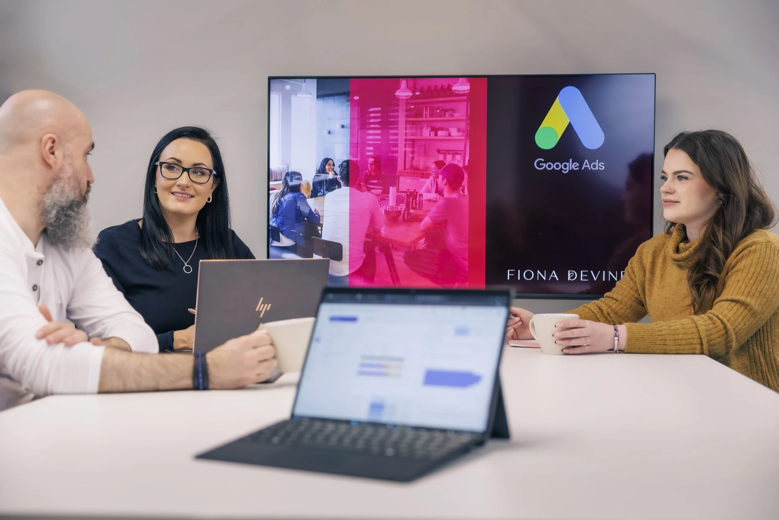 Three people sitting at a conference table with laptops, cups, and a large screen behind them displaying Google Ads and a meeting. One woman is smiling and looking at a man with a beard, and another woman is looking at the man, holding a cup.