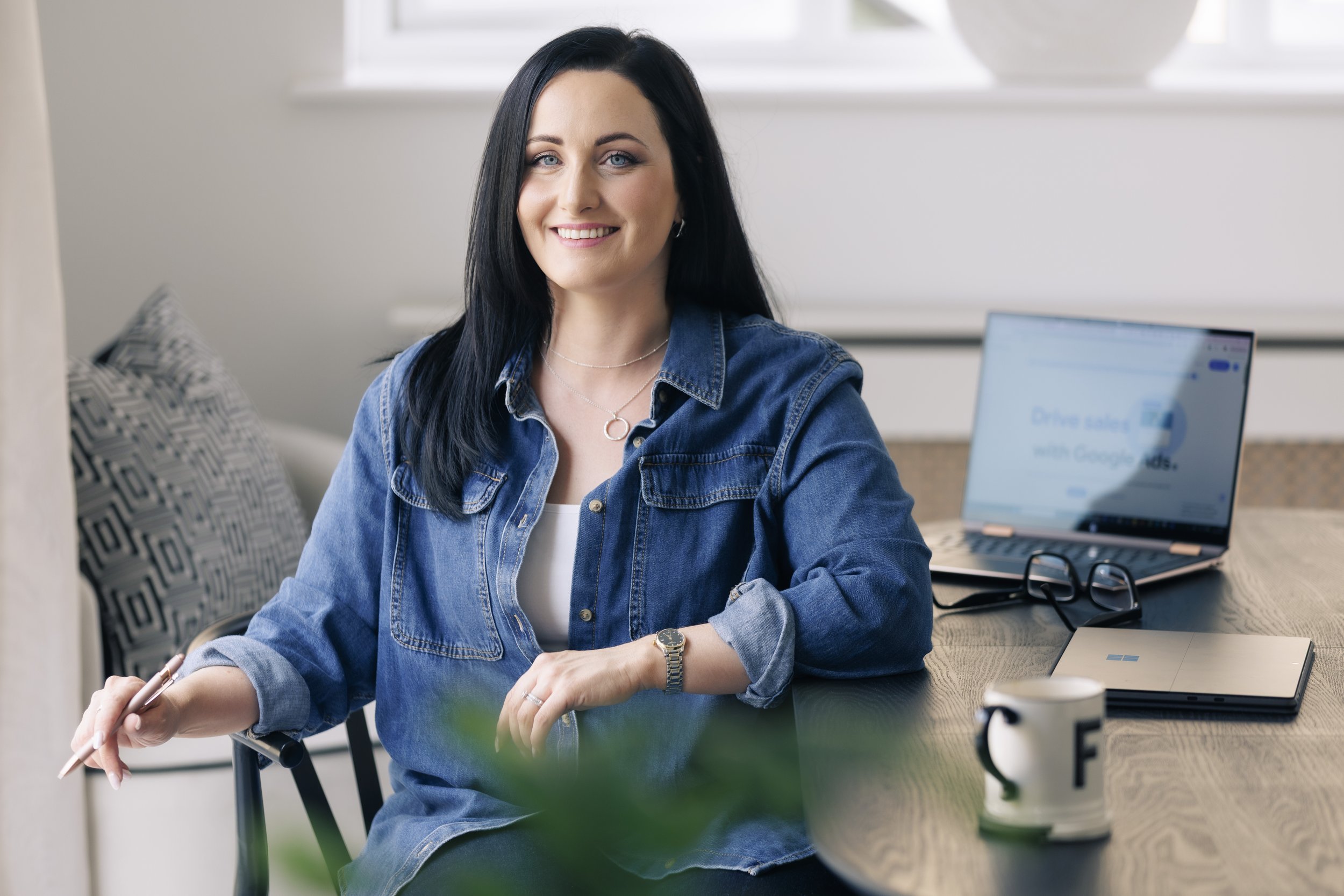 A woman with black hair smiling and seated at a desk, wearing a denim jacket, with a laptop, glasses, and coffee mug nearby.