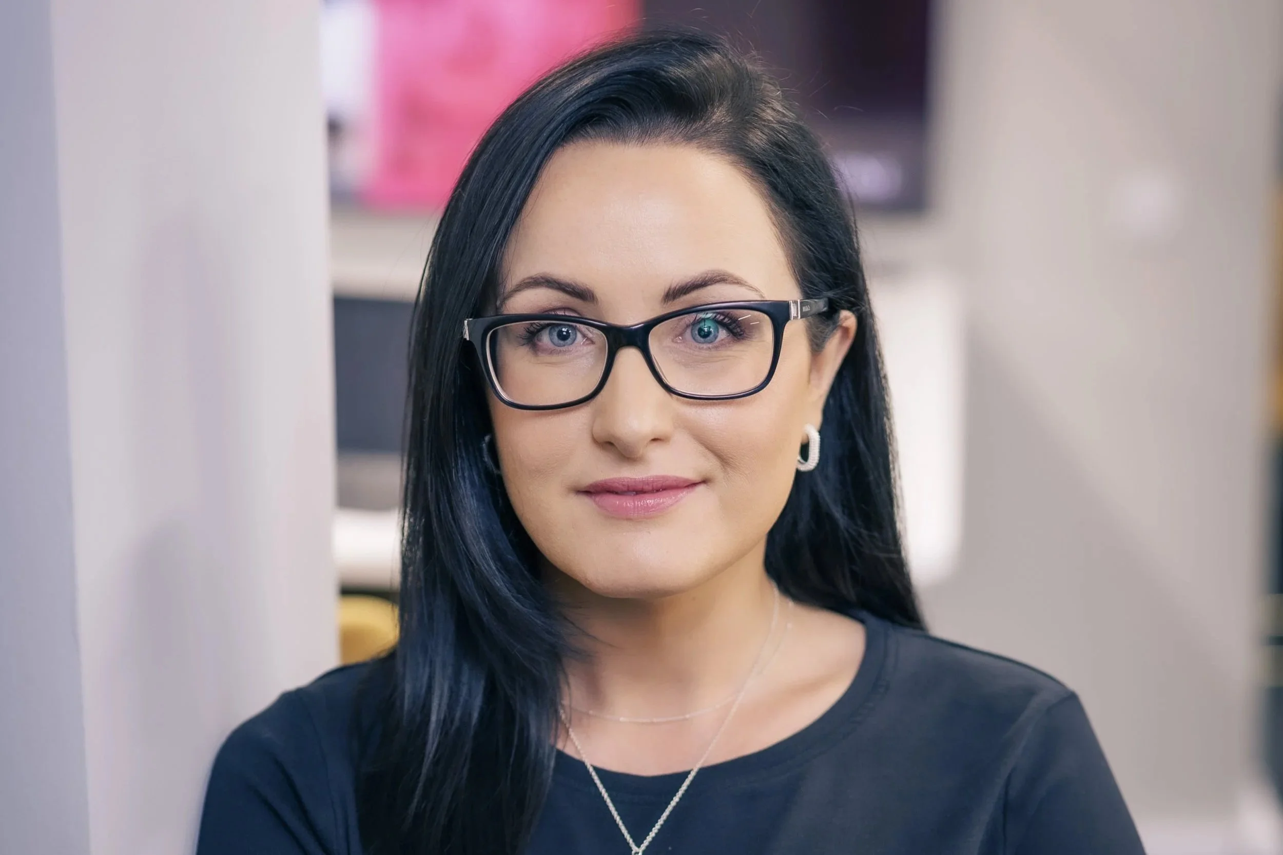 A woman with black hair and blue eyes wearing black glasses and a black shirt, smiling in an indoor setting.