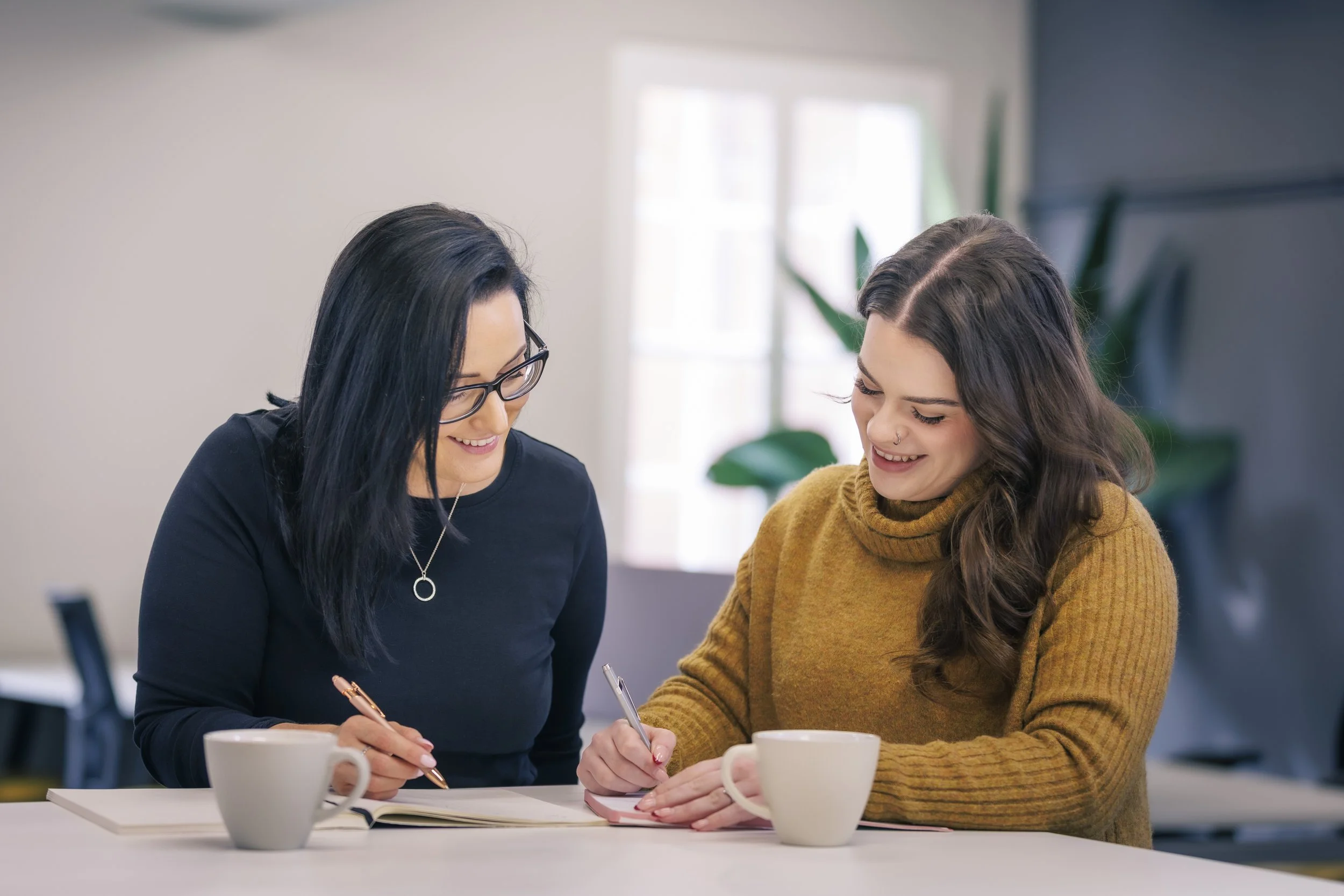 Two women sitting at a white table, smiling and writing in notebooks, with two white coffee mugs in front of them, in a well-lit room with a large window and green plant in the background.