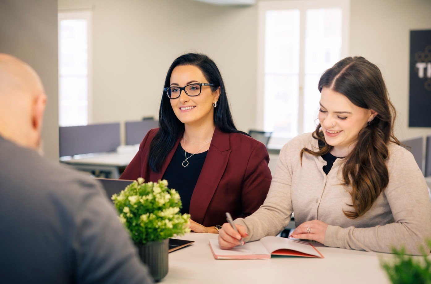 Two women and a man sitting at a table in a modern office, engaged in a discussion. The woman on the right is writing in a notebook while the woman on the left is smiling, wearing glasses and a maroon blazer. There is a potted plant on the table.