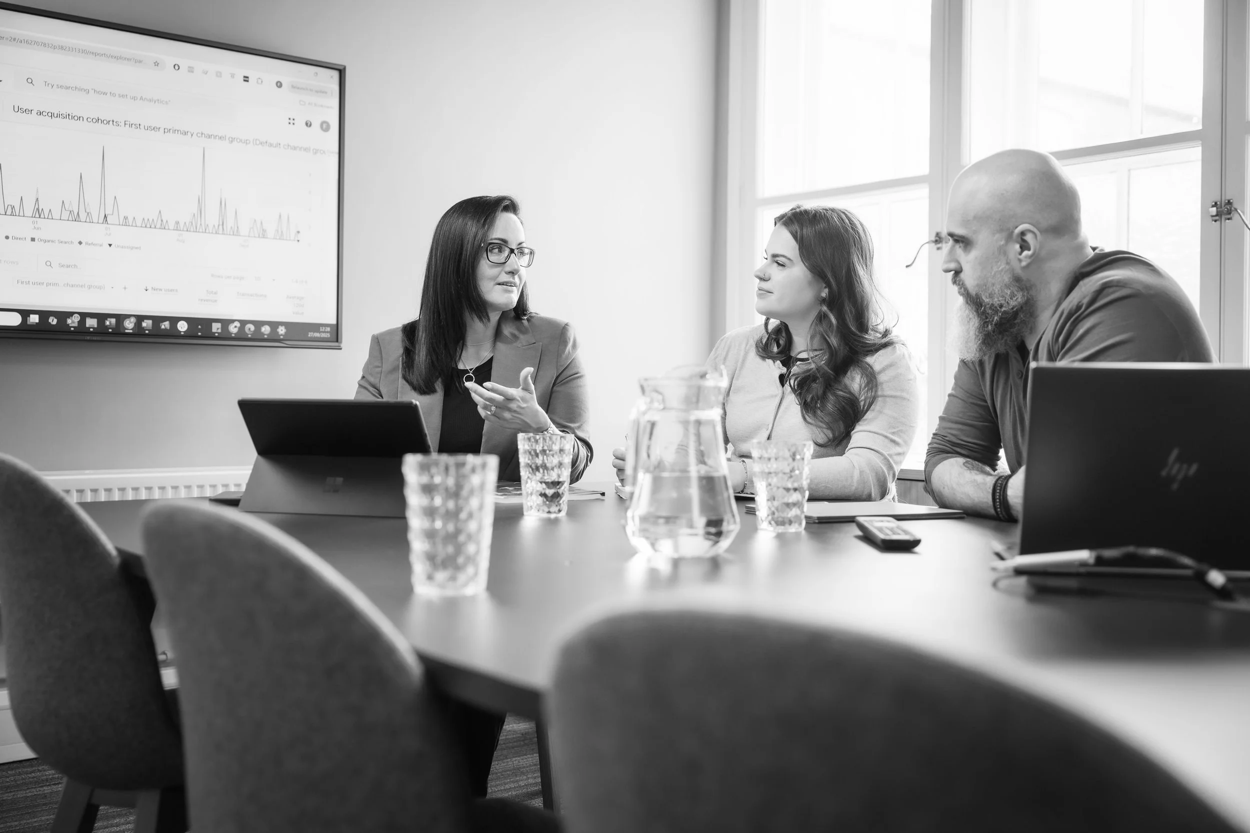 Three people sitting at a conference table in a meeting room, engaged in discussion with a large screen displaying a graph. The group includes a woman with glasses speaking, a woman with long hair listening, and a man with a beard and bald head listening. There are glasses and a pitcher of water on the table.