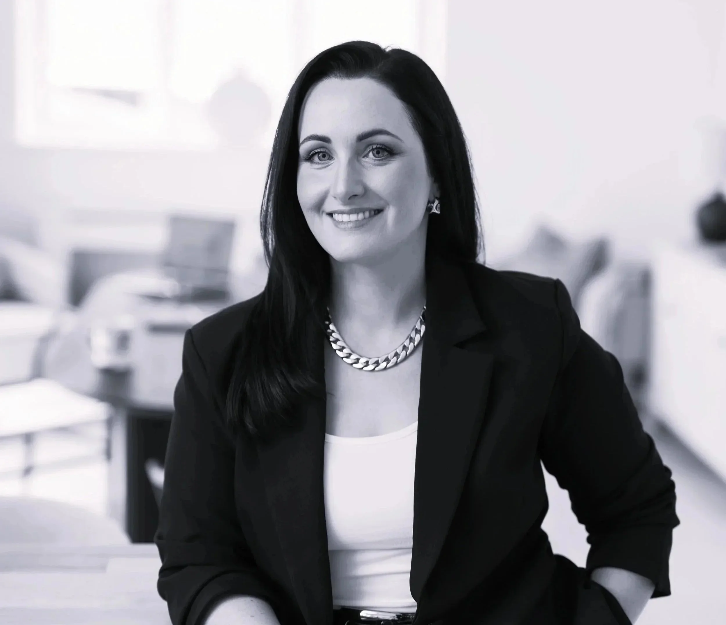 A woman with dark hair, light skin, wearing a black blazer, white top, chain necklace, and earrings, smiling in an office setting.