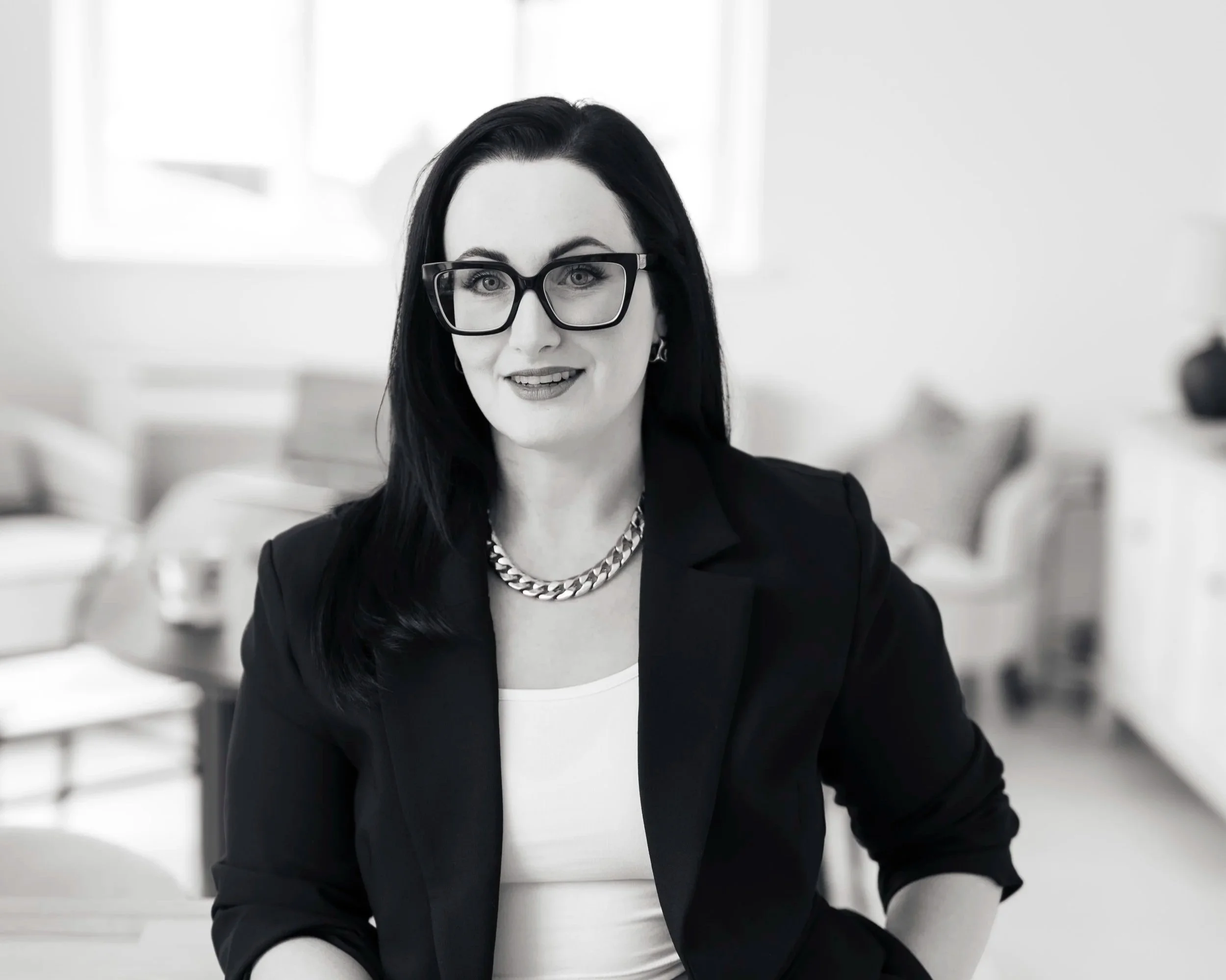 Black and white photo of a woman with long dark hair, wearing glasses, a blazer, and a chain necklace, sitting in a bright room with modern furniture.