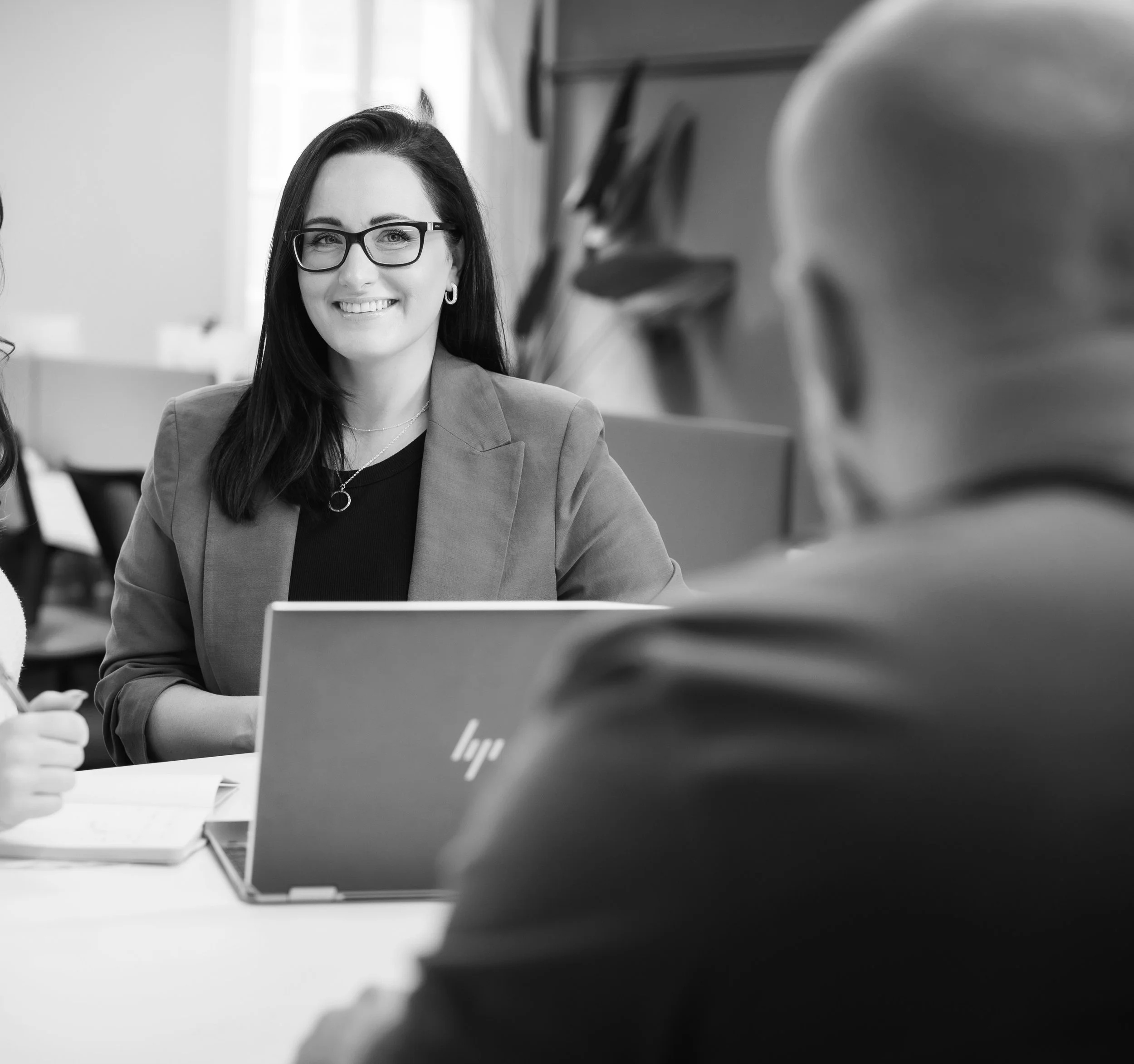 A woman with dark hair, glasses, and a blazer smiling during a business meeting.