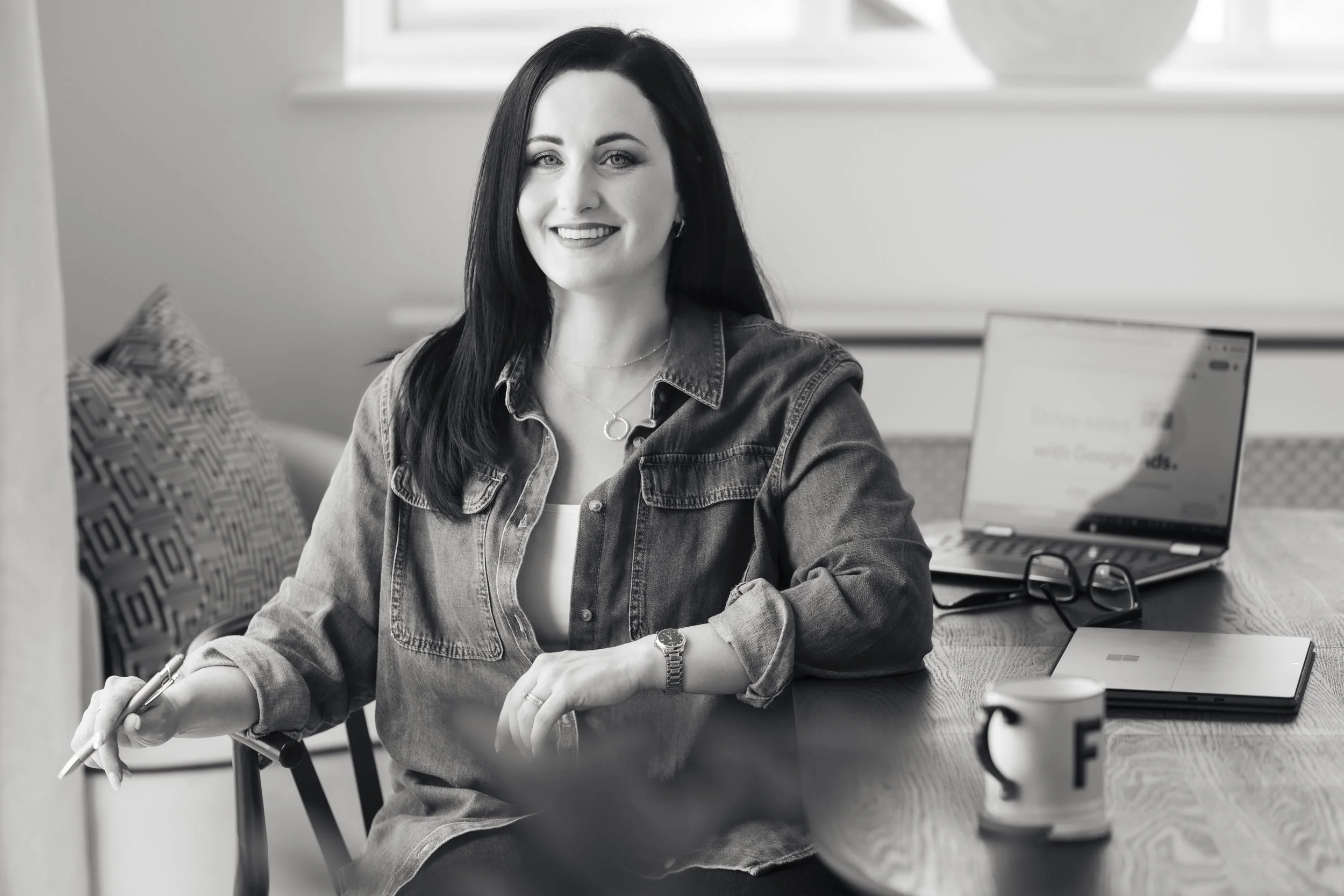A woman with long dark hair sitting at a table with a laptop, a cup, and a pair of glasses, smiling and holding a pen in her hand.