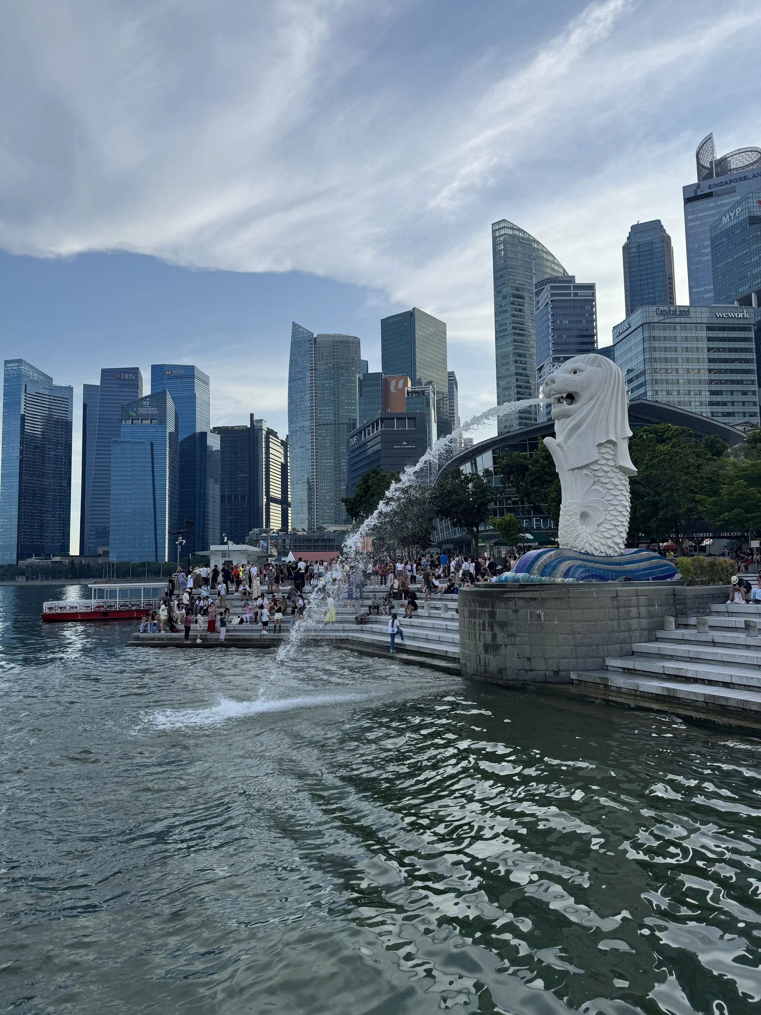 The image features the Merlion statue spouting water with a cityscape of modern skyscrapers in the background, crowded with people along the waterfront.