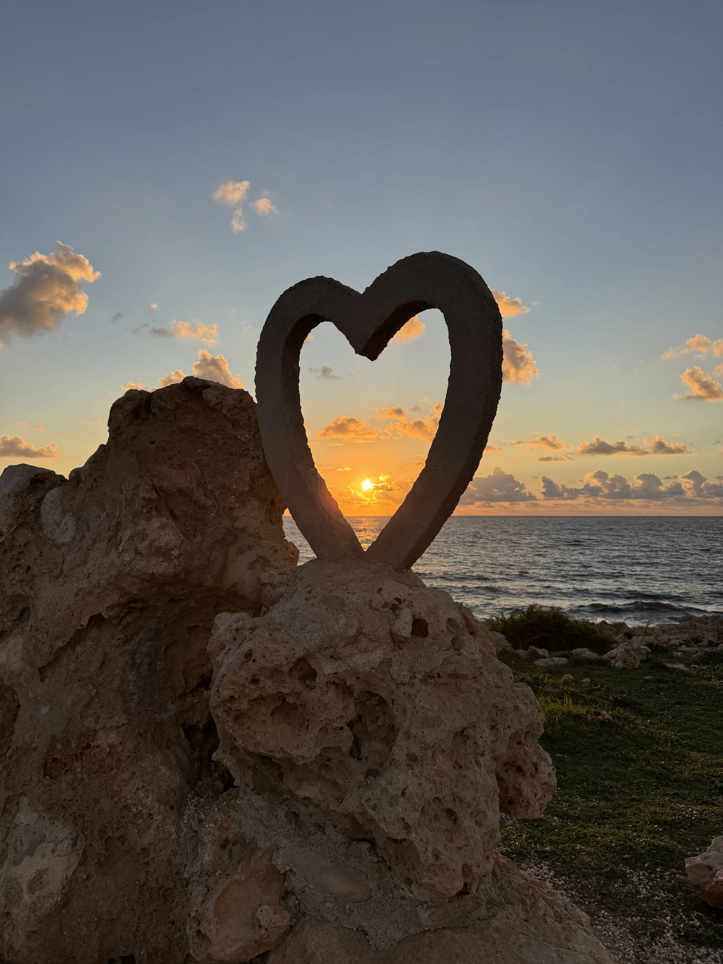 Sunset over the ocean seen from a rocky coastline with a large heart-shaped sculpture in foreground.