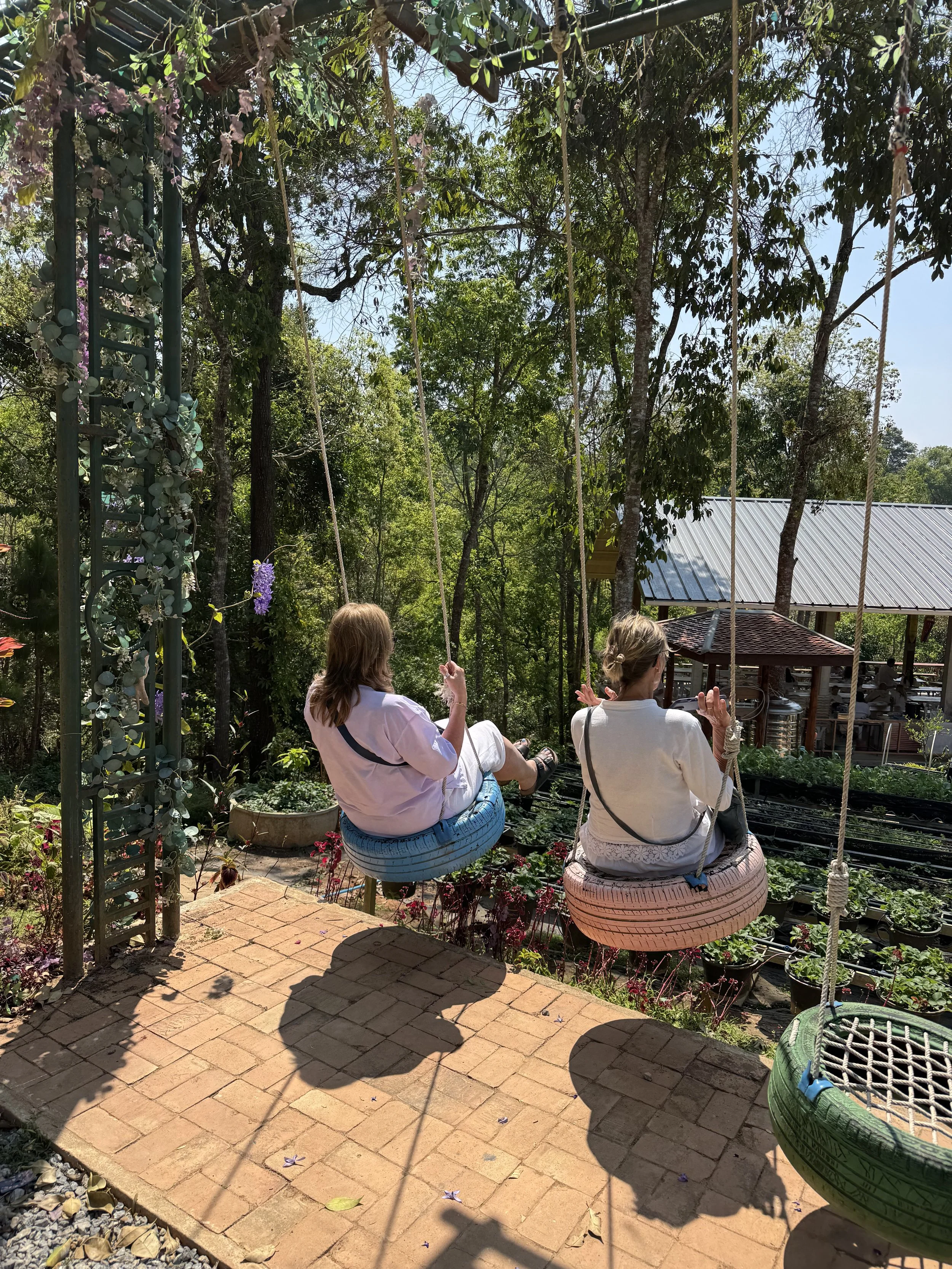 Two women sitting on tire swings in an outdoor garden area with lush green trees, plants, and a metal-roofed building in the background.