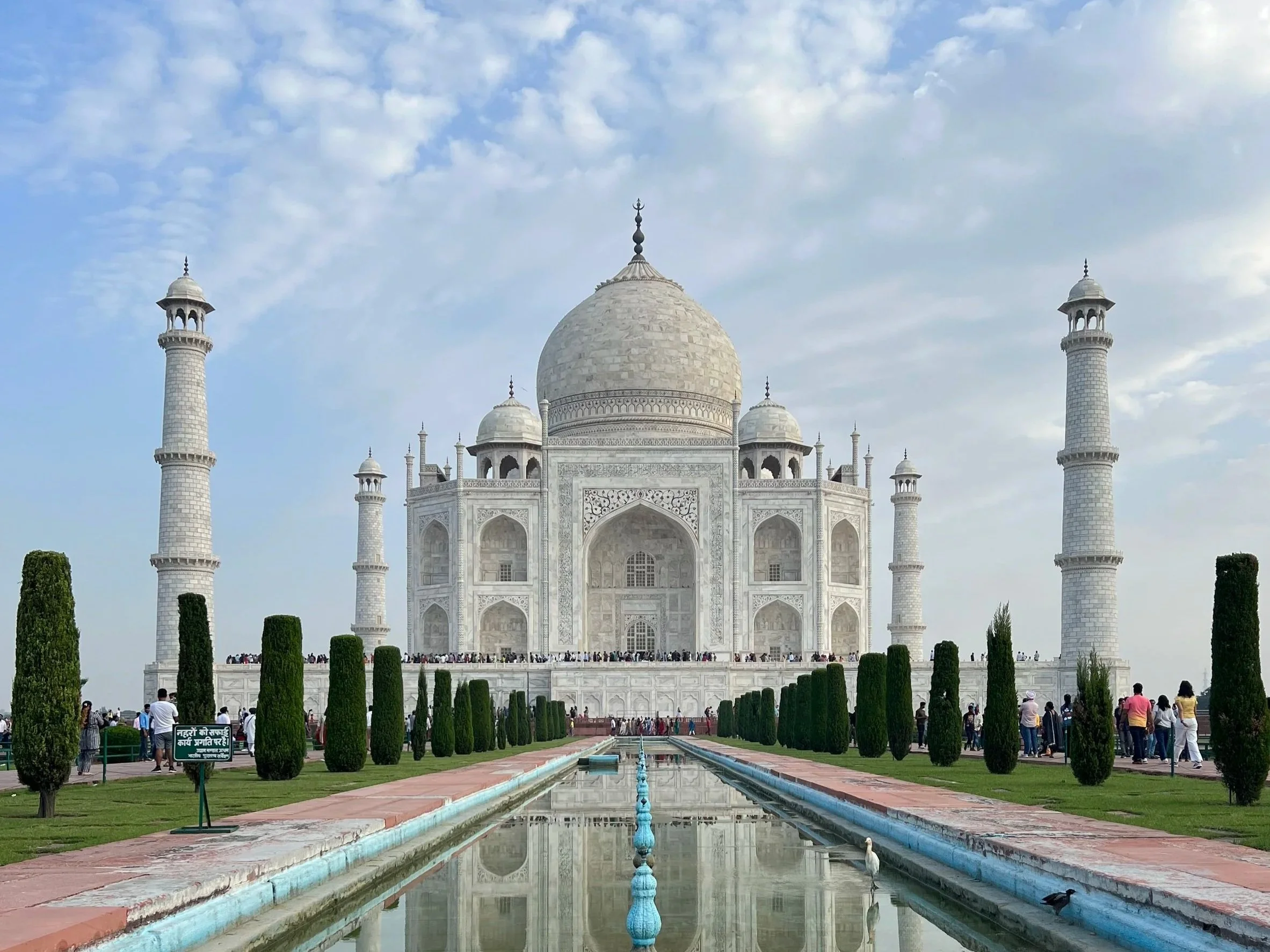 The Taj Mahal, a white marble mausoleum with a large central dome and four minarets, surrounded by green lawns, trees, and a reflecting pool with birds, under a partly cloudy sky.